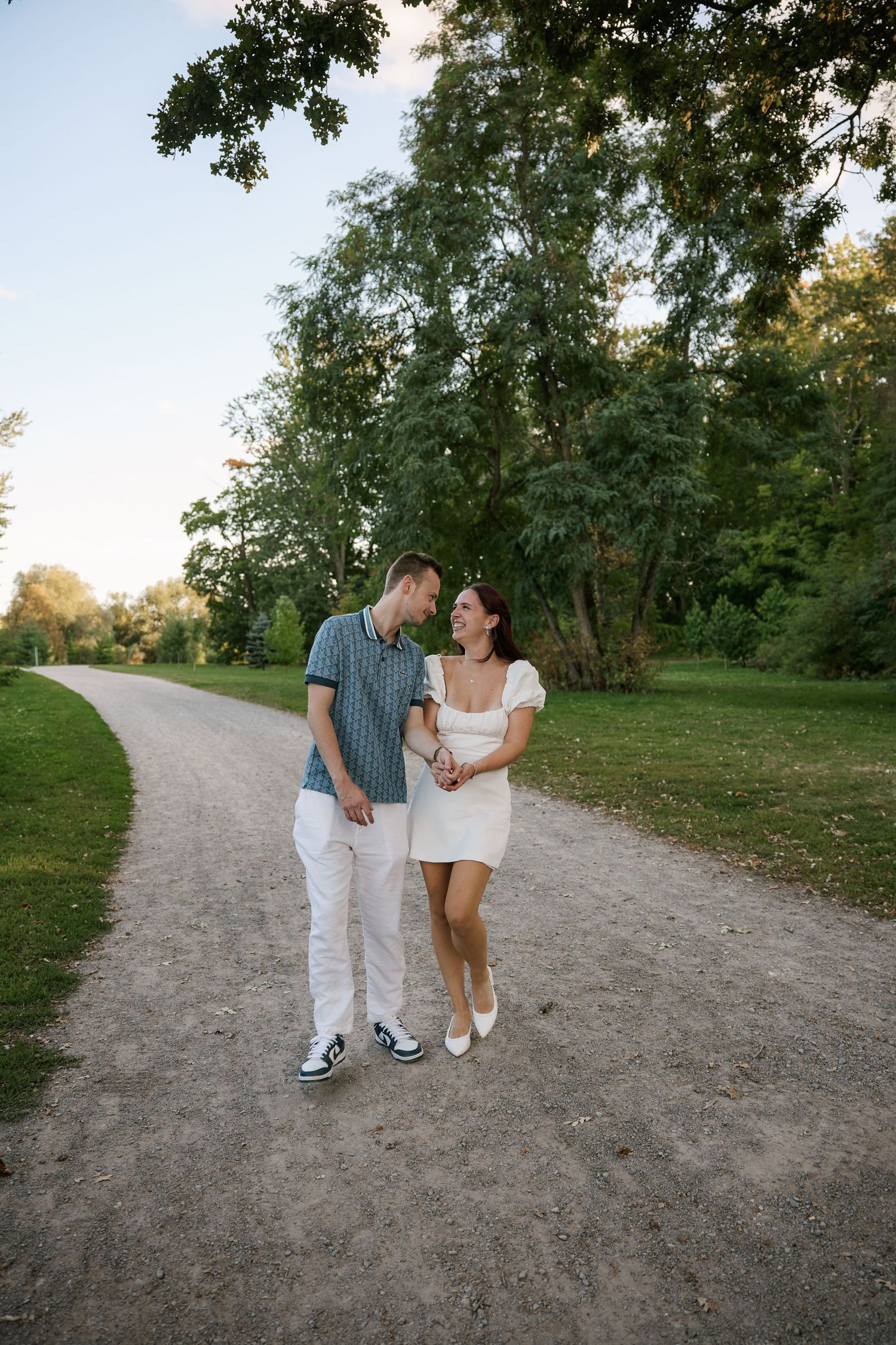Couple holding hands and smiling at each other while walking on a park path surrounded by lush green trees.