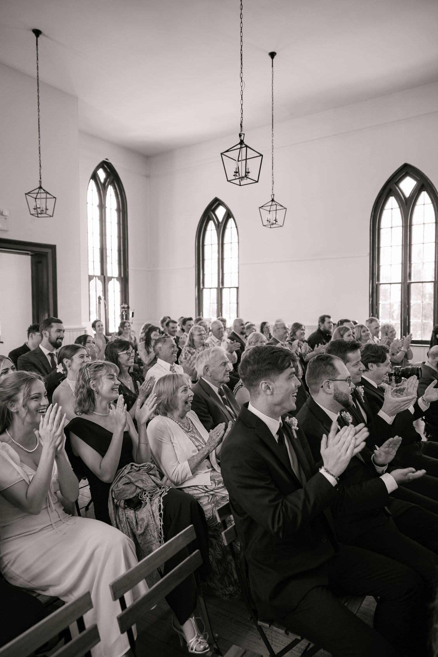 Black and white photo of wedding guests clapping in a chapel with arched windows and pendant lights.