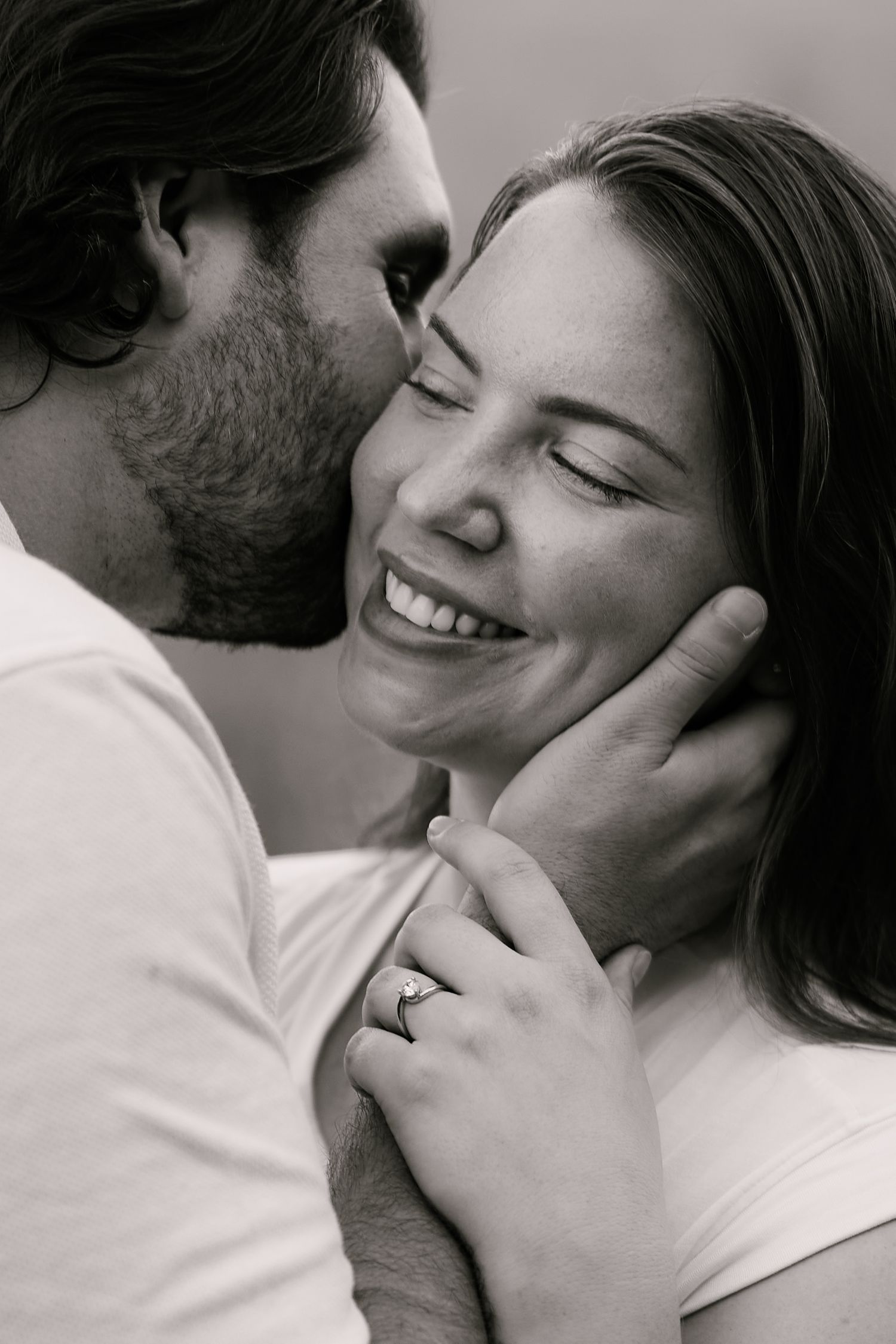 Black and white photo of a smiling woman with engagement ring being kissed on cheek by bearded man.