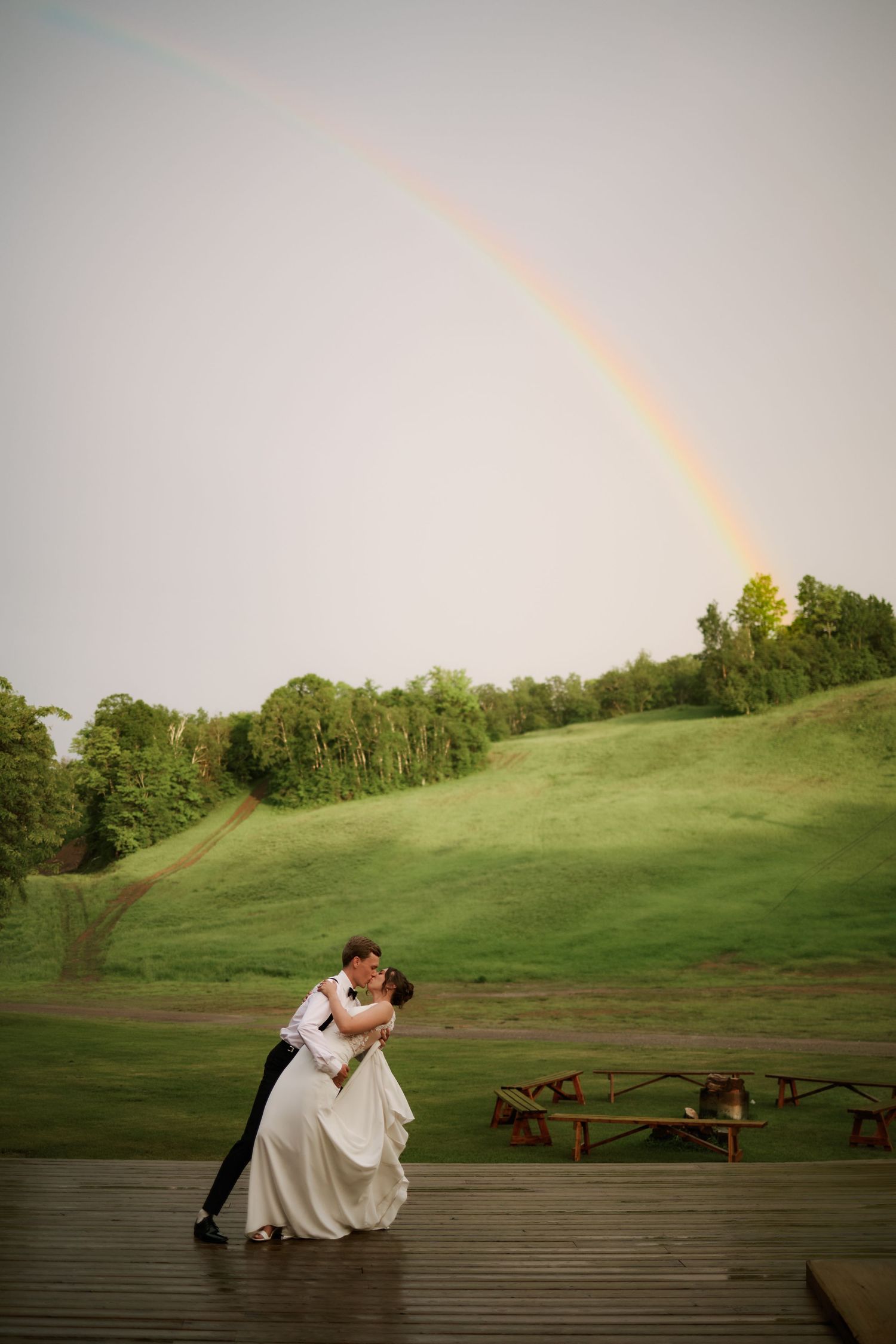 Bride and groom share a romantic dip kiss on a wooden deck with a stunning rainbow arching over green hills.