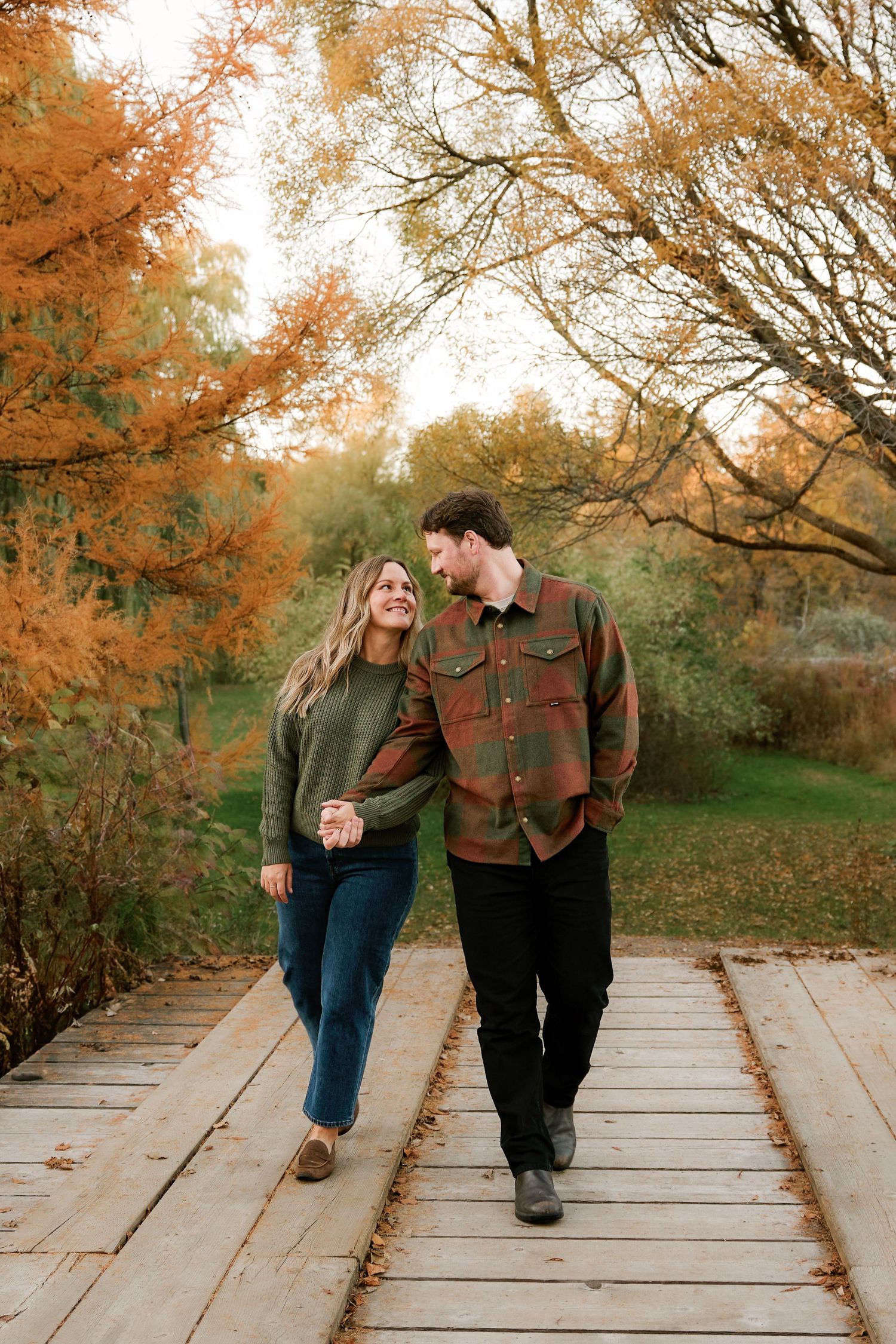 A couple walking hand in hand on a wooden boardwalk surrounded by autumn foliage.