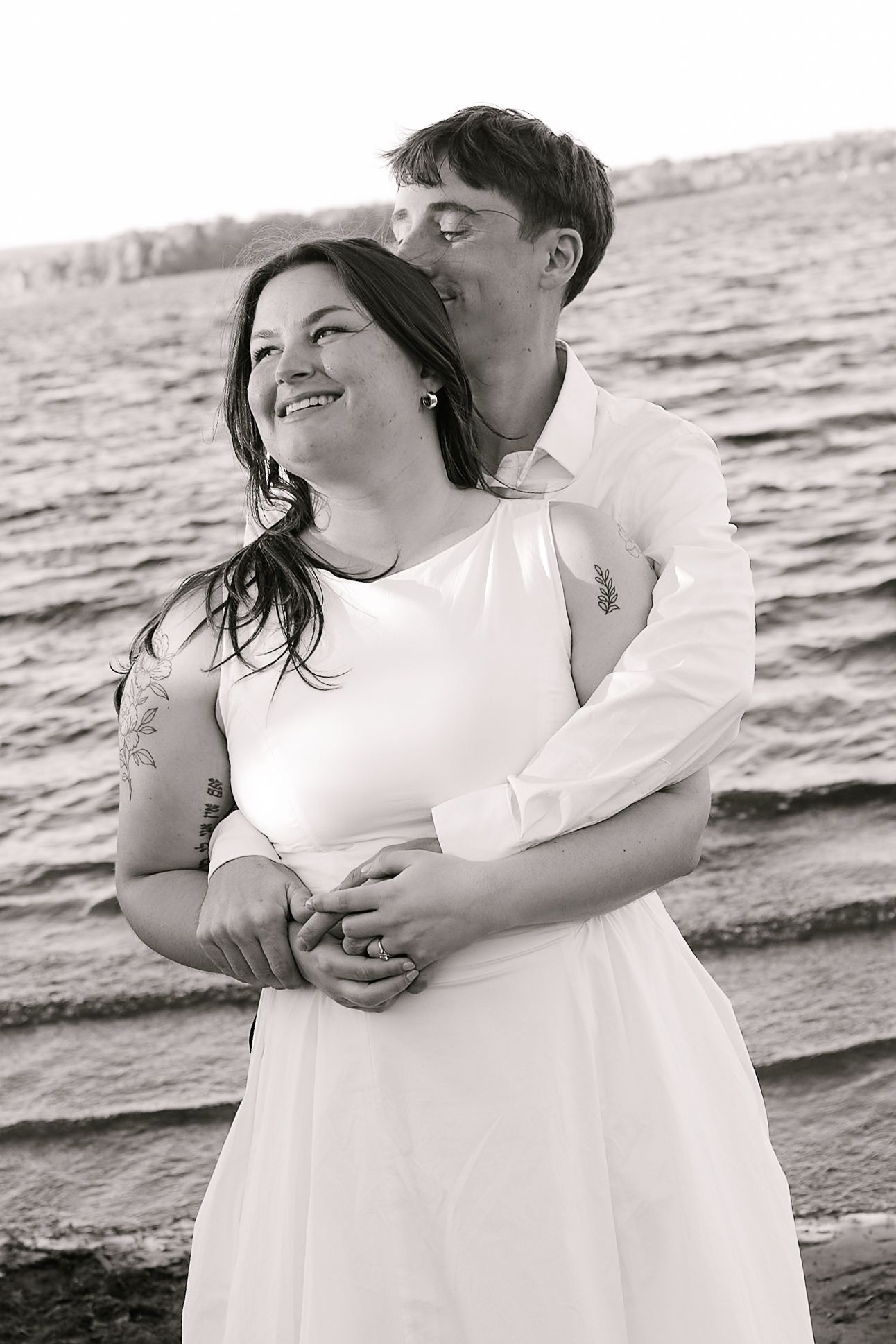 Black and white photo of a couple embracing on a beach, woman in white dress smiling as man kisses her head.