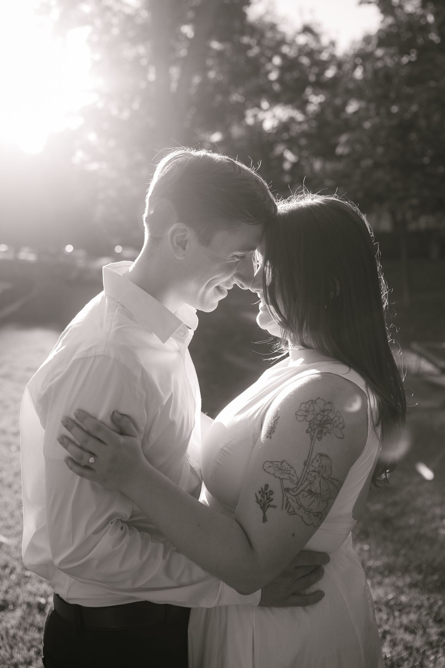 Black and white photo of a couple embracing and smiling at each other outdoors, woman with floral arm tattoo.
