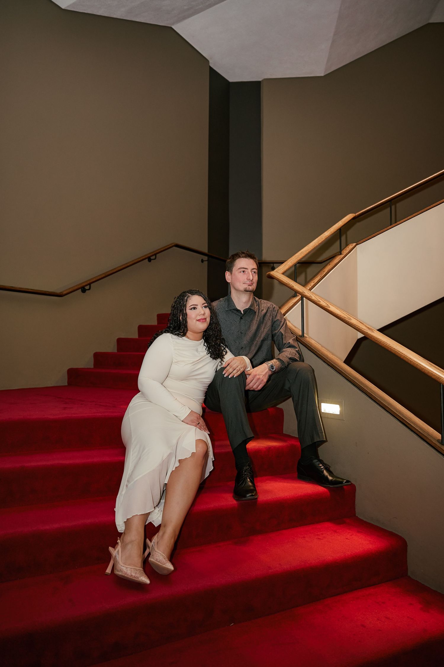 Couple in elegant attire sitting on red carpeted stairs, woman in white dress and man in gray suit, gazing upward.