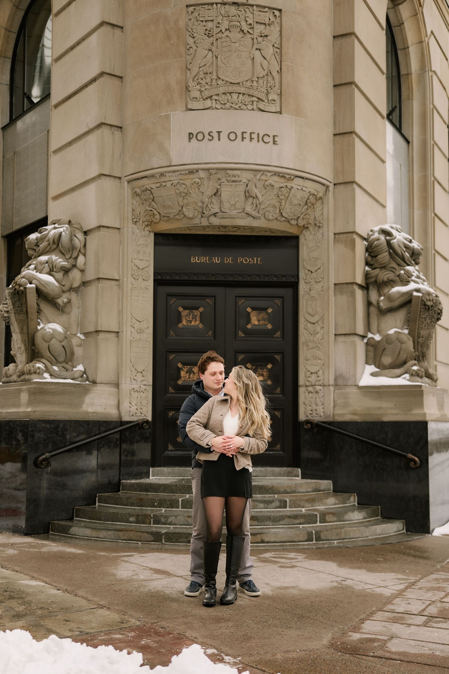Couple embracing in front of a historic post office building with stone statues and ornate architecture in winter.