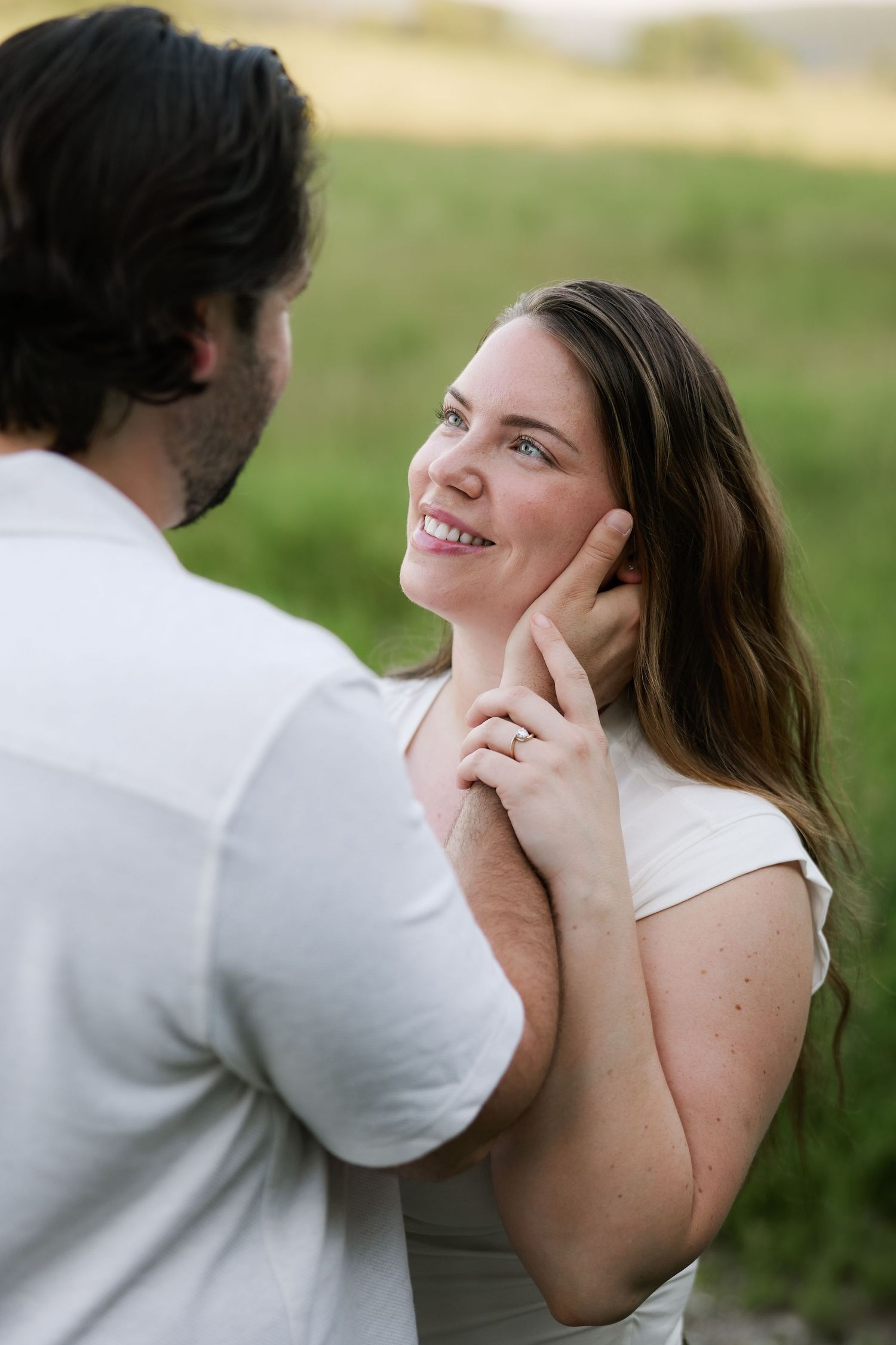 A smiling woman gazes at a man outdoors, wearing an engagement ring, with green fields in the background.