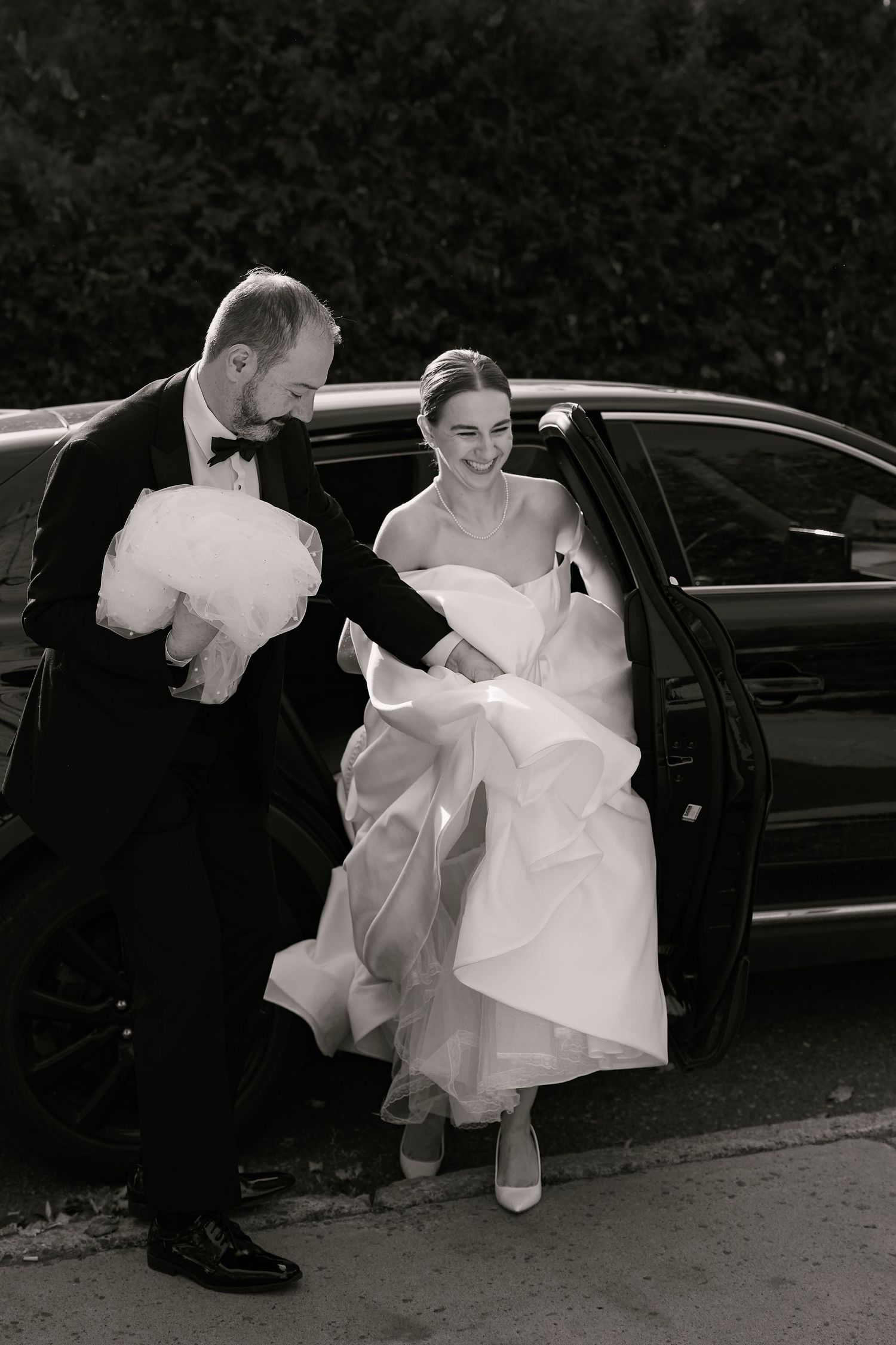 A couple in wedding attire getting out of a black car.