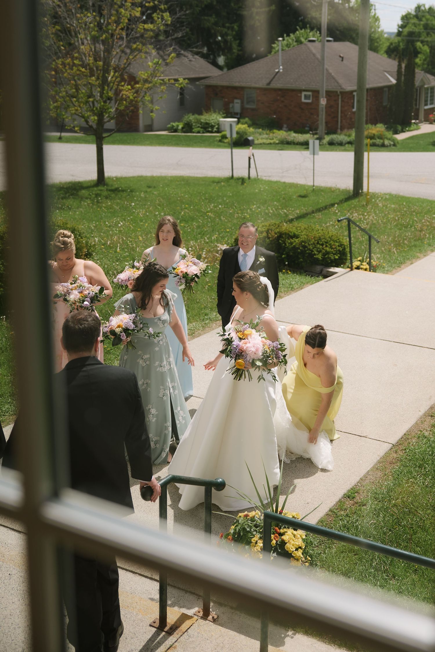 Bride in white gown with bridesmaids in sage and yellow dresses ascending church steps holding colorful bouquets.