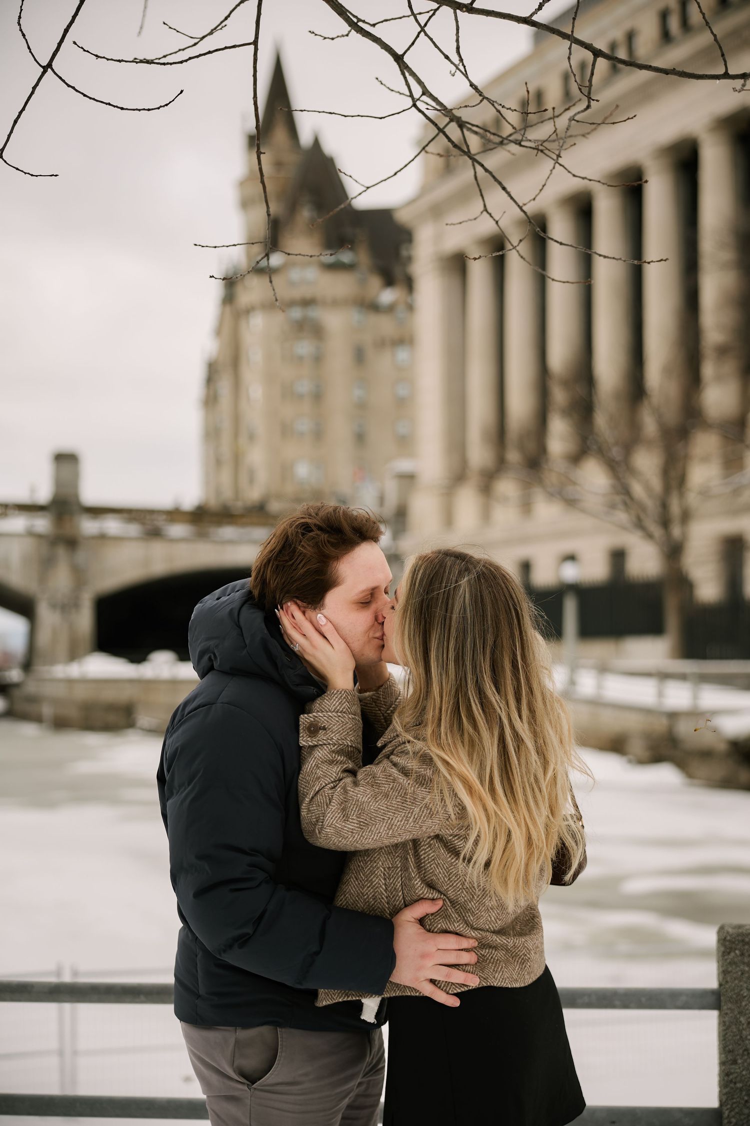 Couple sharing a romantic kiss outdoors in winter, embracing near a snowy riverbank with a historic building backdrop.