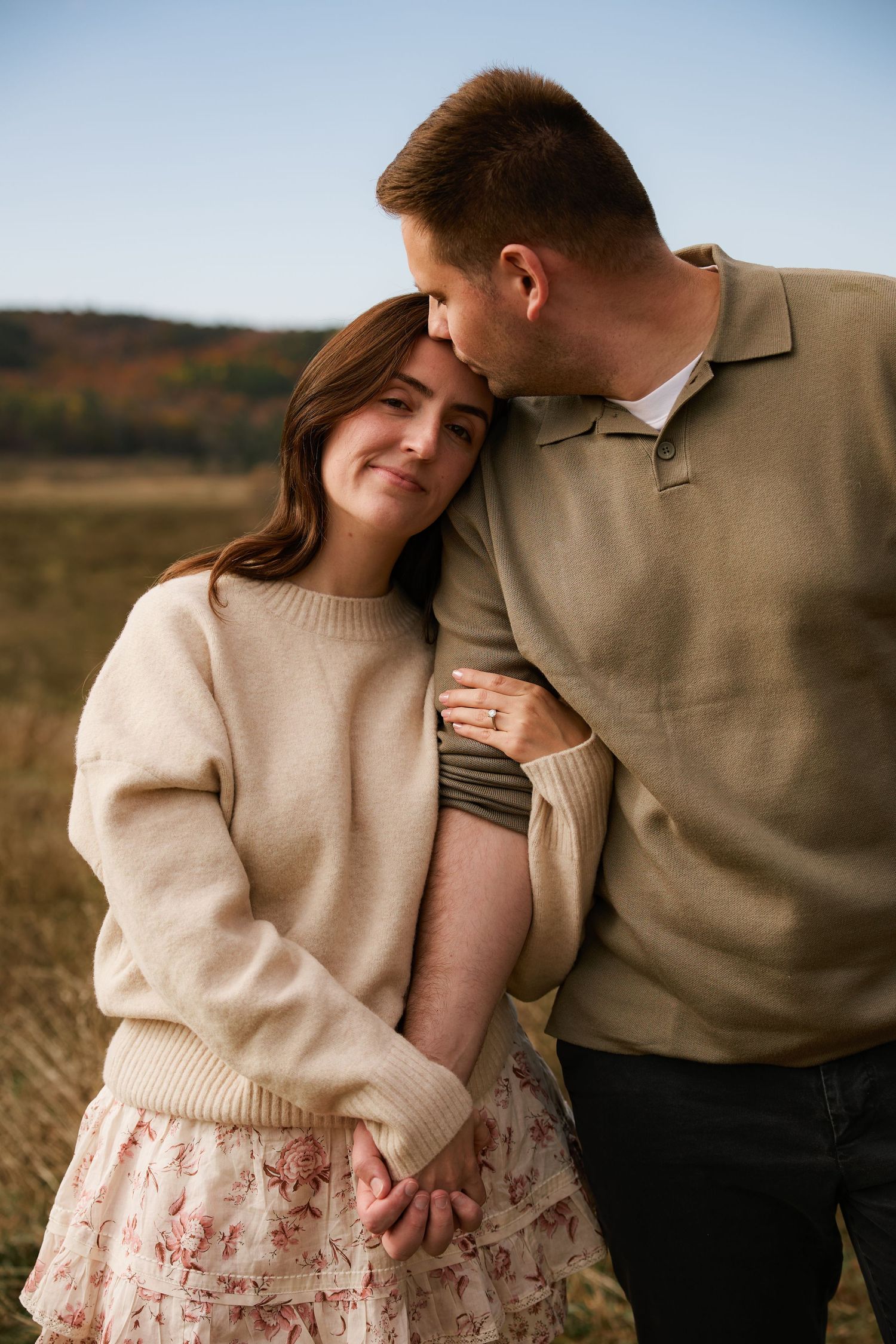 Couple embracing outdoors in autumn field, woman in floral skirt and cream sweater leans on man kissing her head.