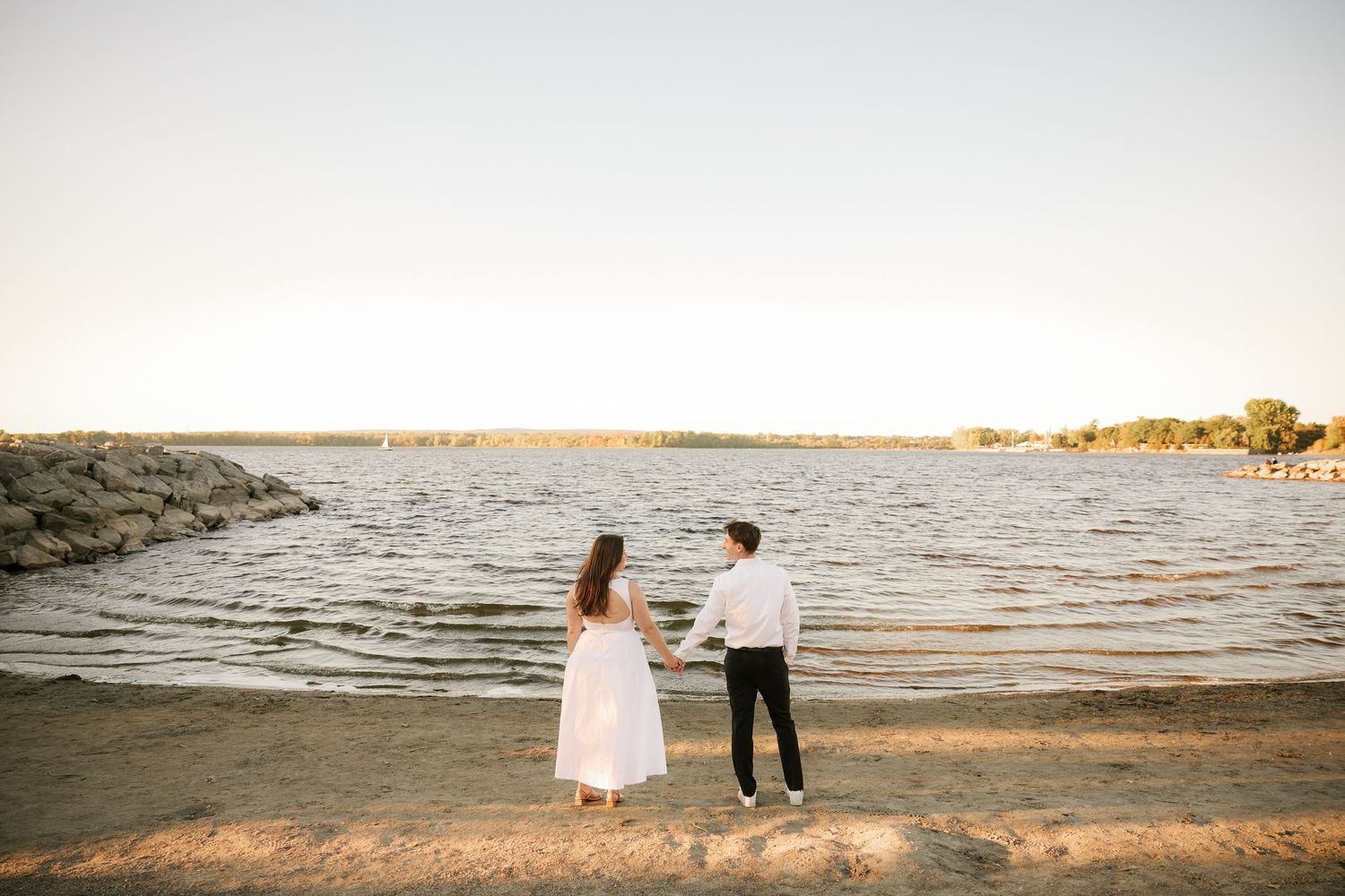 Couple holding hands on sandy beach shore at sunset, woman in white dress and man in white shirt near calm water.