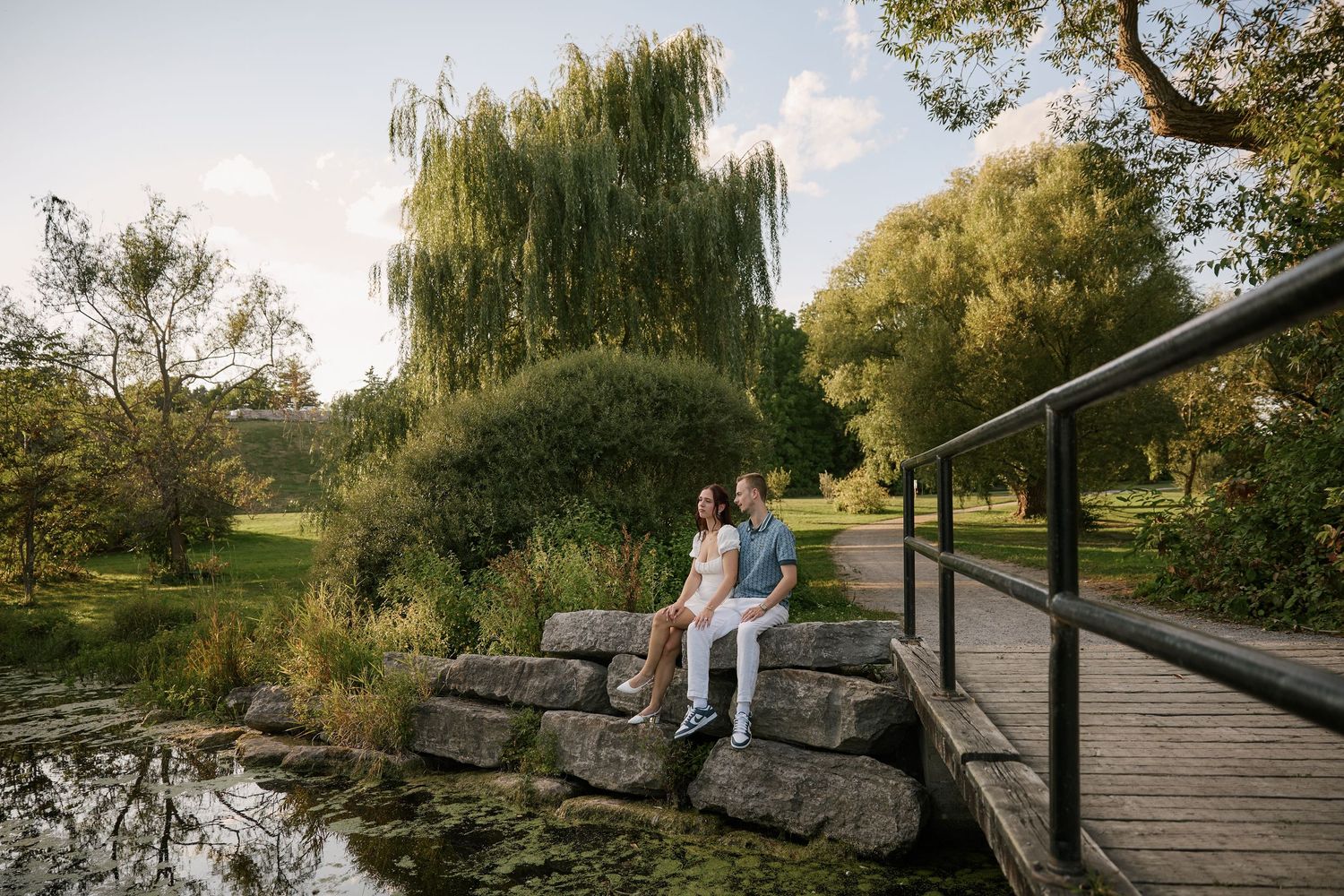 Couple sitting on rocks by a pond in a lush green park near a wooden bridge on a sunny day.