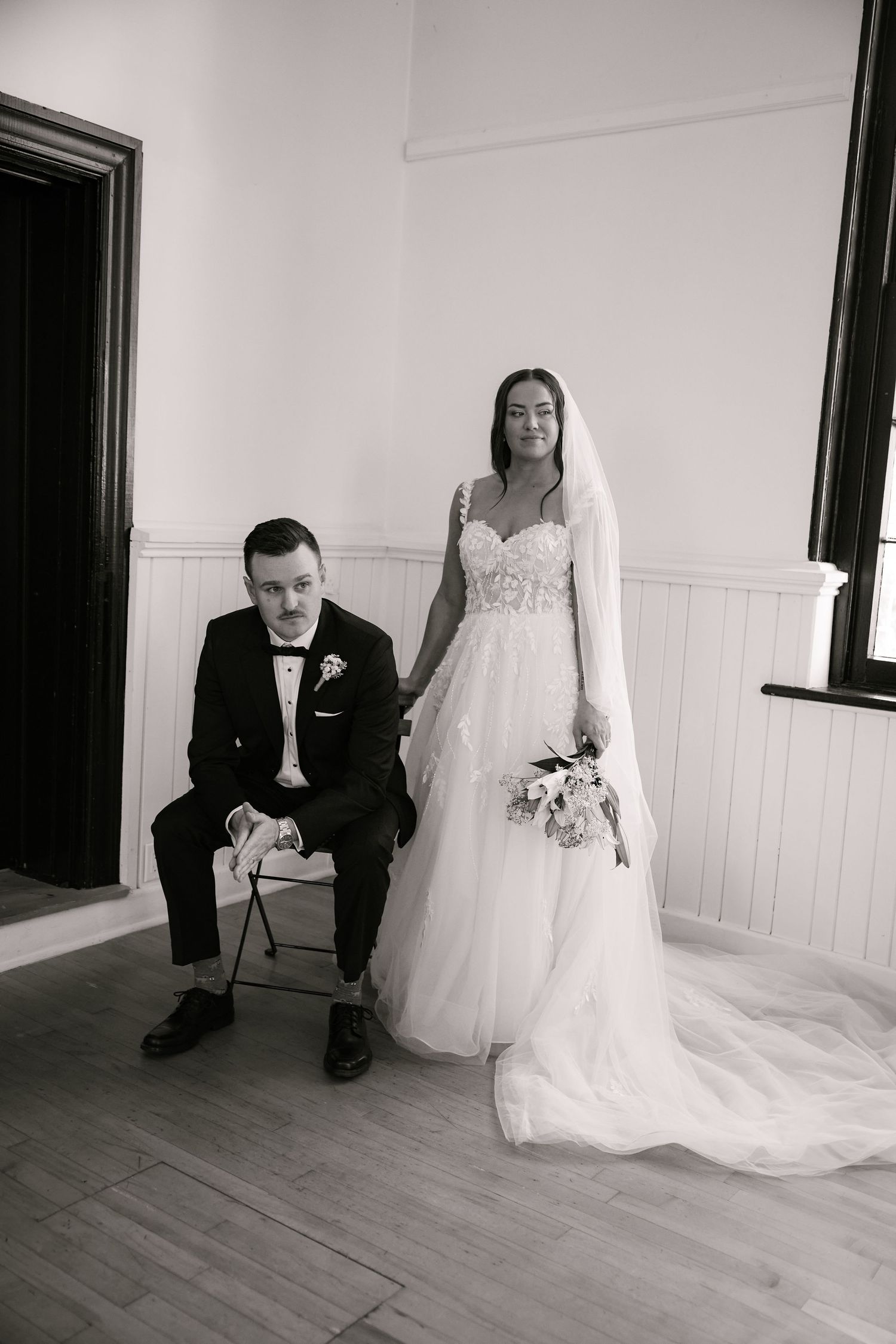Black and white wedding portrait of bride in lace gown holding bouquet standing beside groom in tuxedo seated on chair.