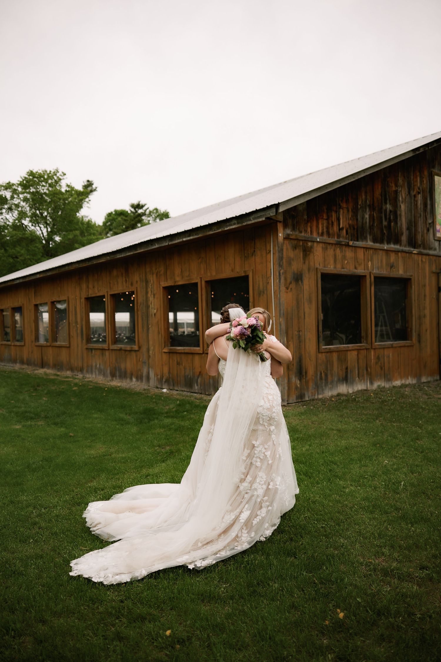Bride in lace wedding gown with long train holds bouquet outside rustic wooden barn venue on green lawn.