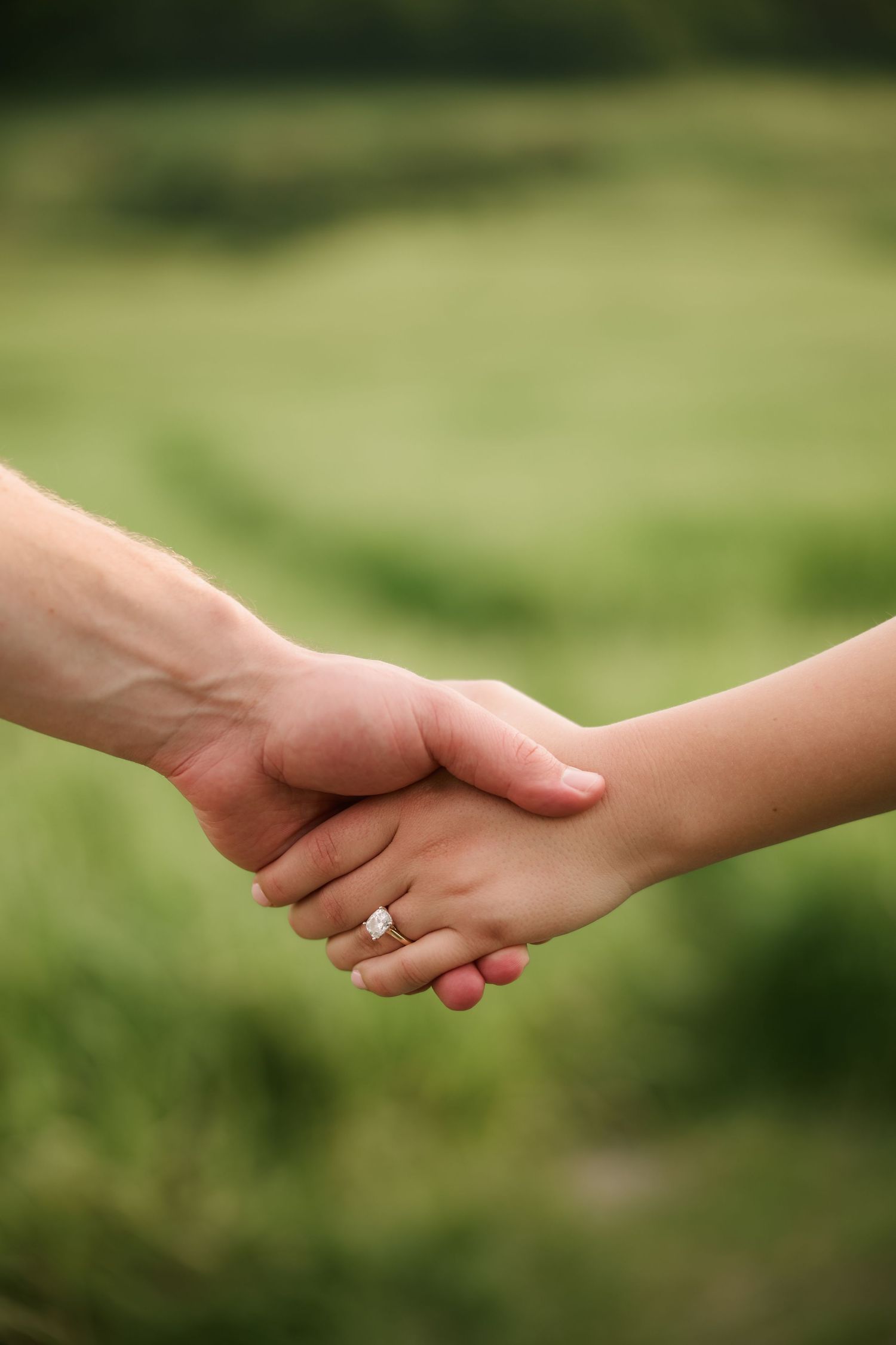 Two people holding hands outdoors with an engagement ring visible, set against a lush green blurred background.