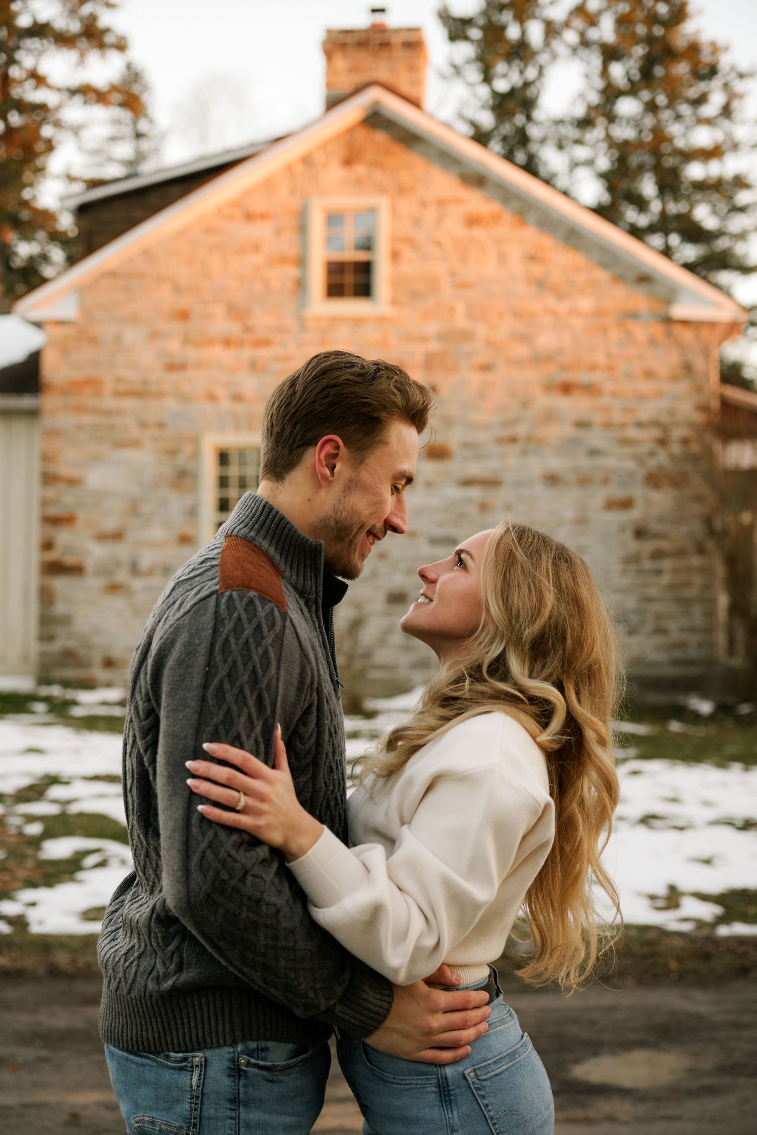 A tender moment shared between a couple in front of a rustic stone house in winter.