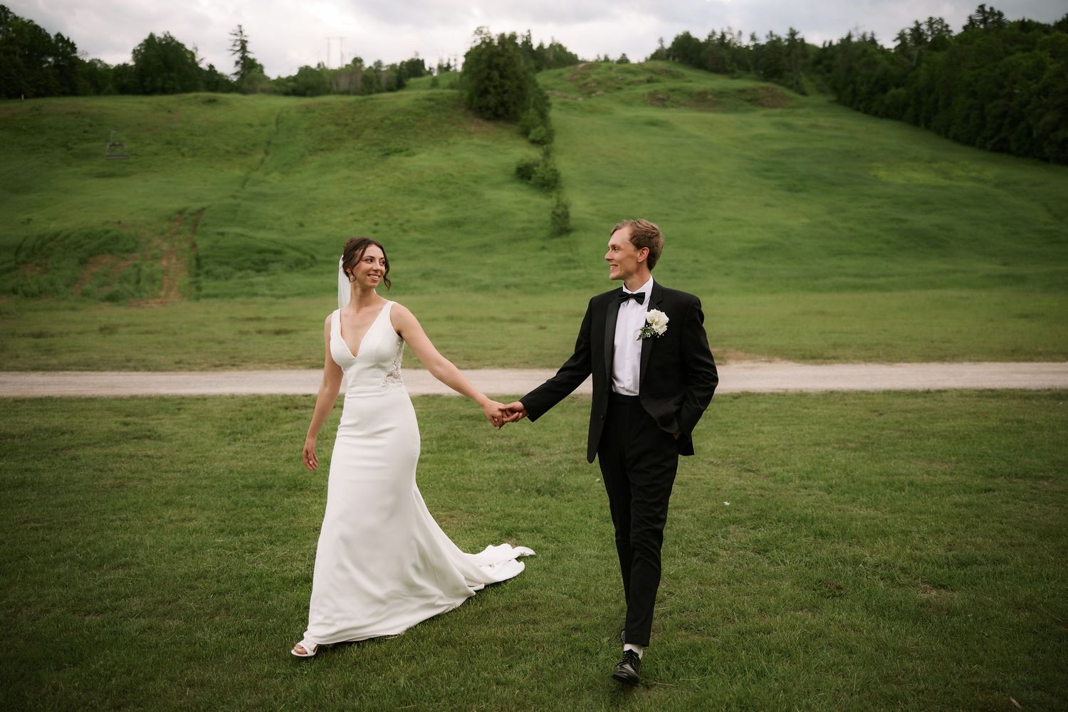 A bride and groom walk hand-in-hand across a lush green hillside on their wedding day.