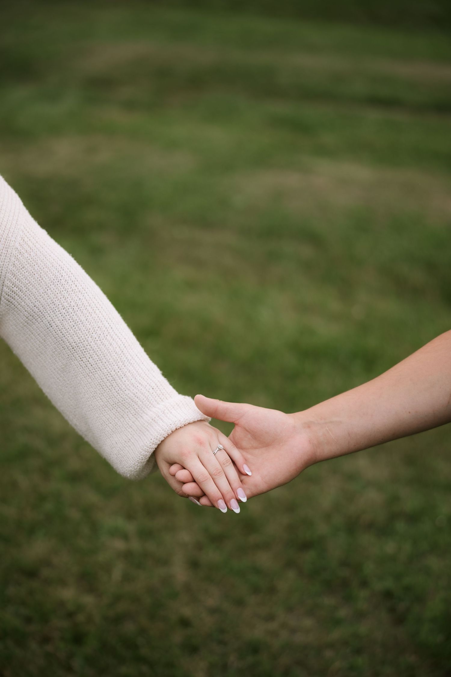 Two people holding hands outdoors on green grass, one wearing a white knit sweater with a delicate ring on their finger.