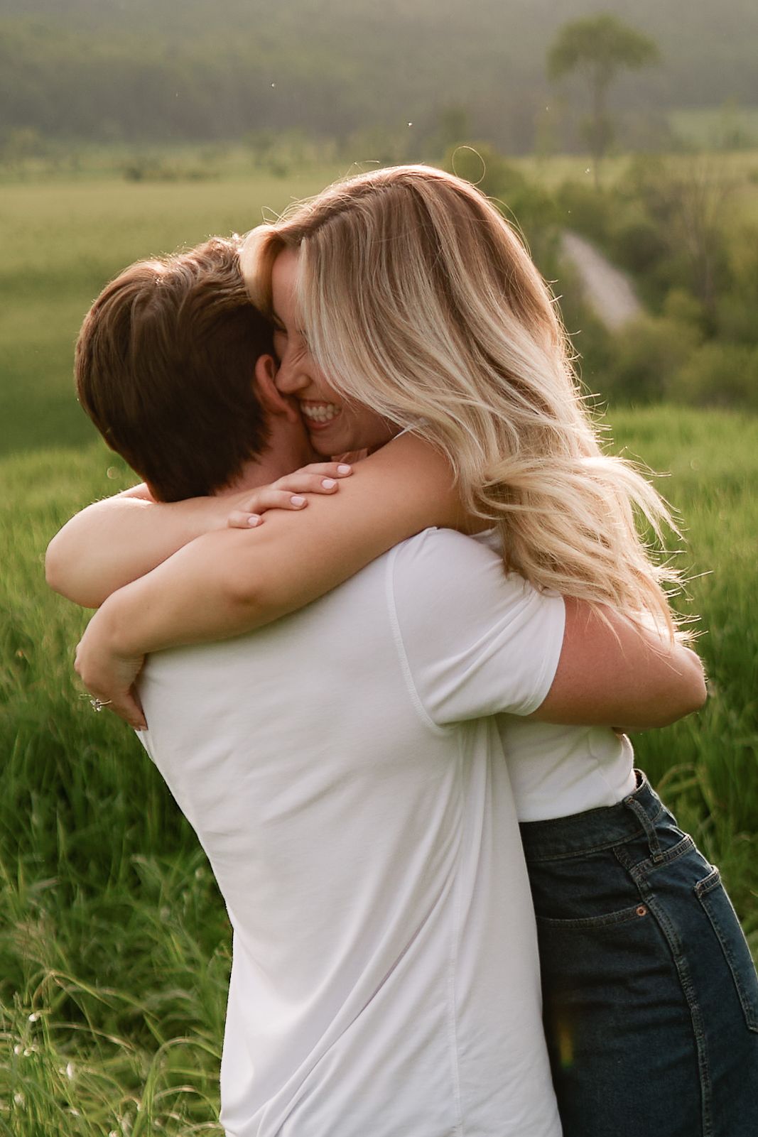 Couple embracing in a green field, woman with blonde hair smiling, both wearing white t-shirts at golden hour.