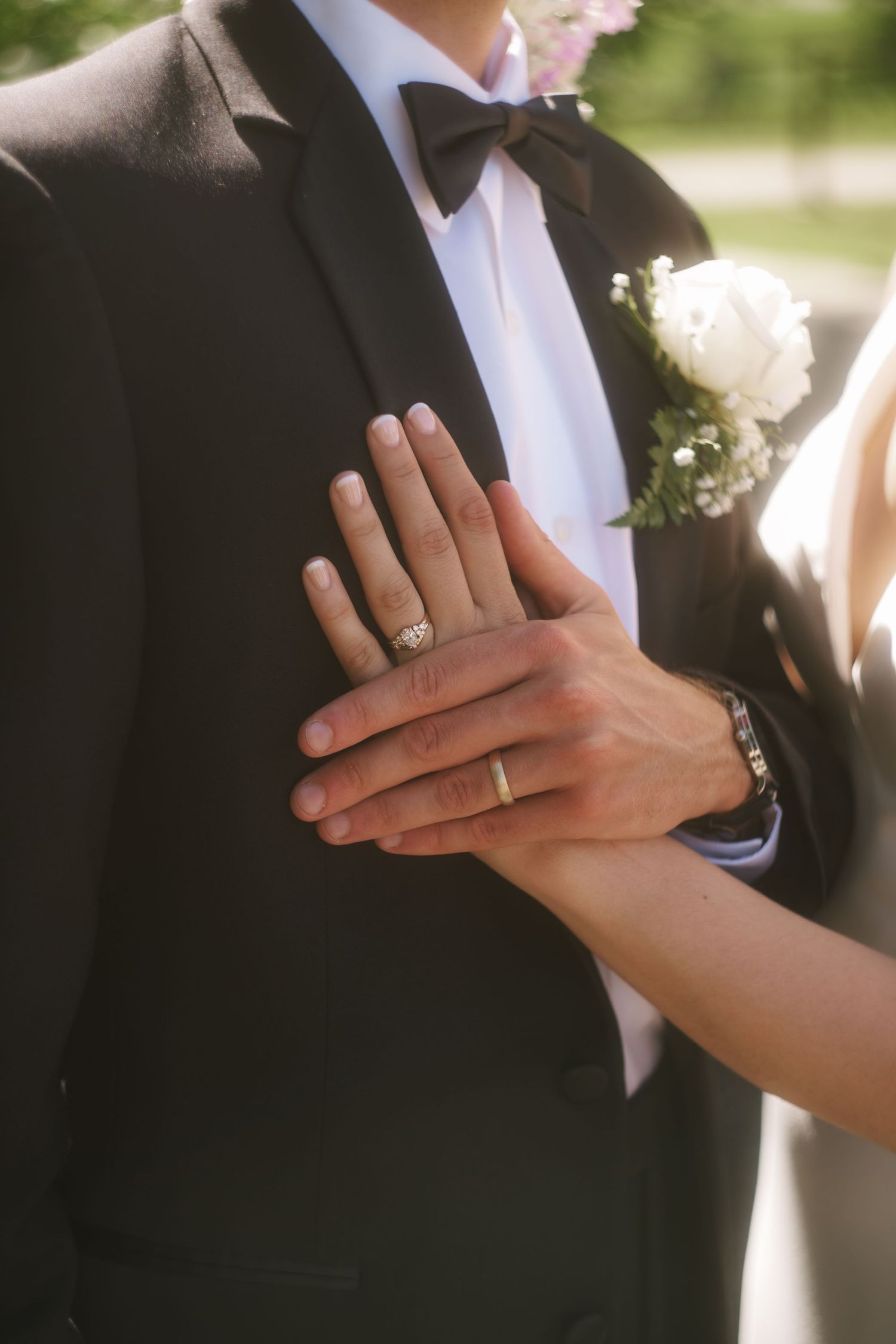 Bride and groom displaying wedding rings, groom in black tuxedo with white boutonniere, bride's hand resting on his chest.