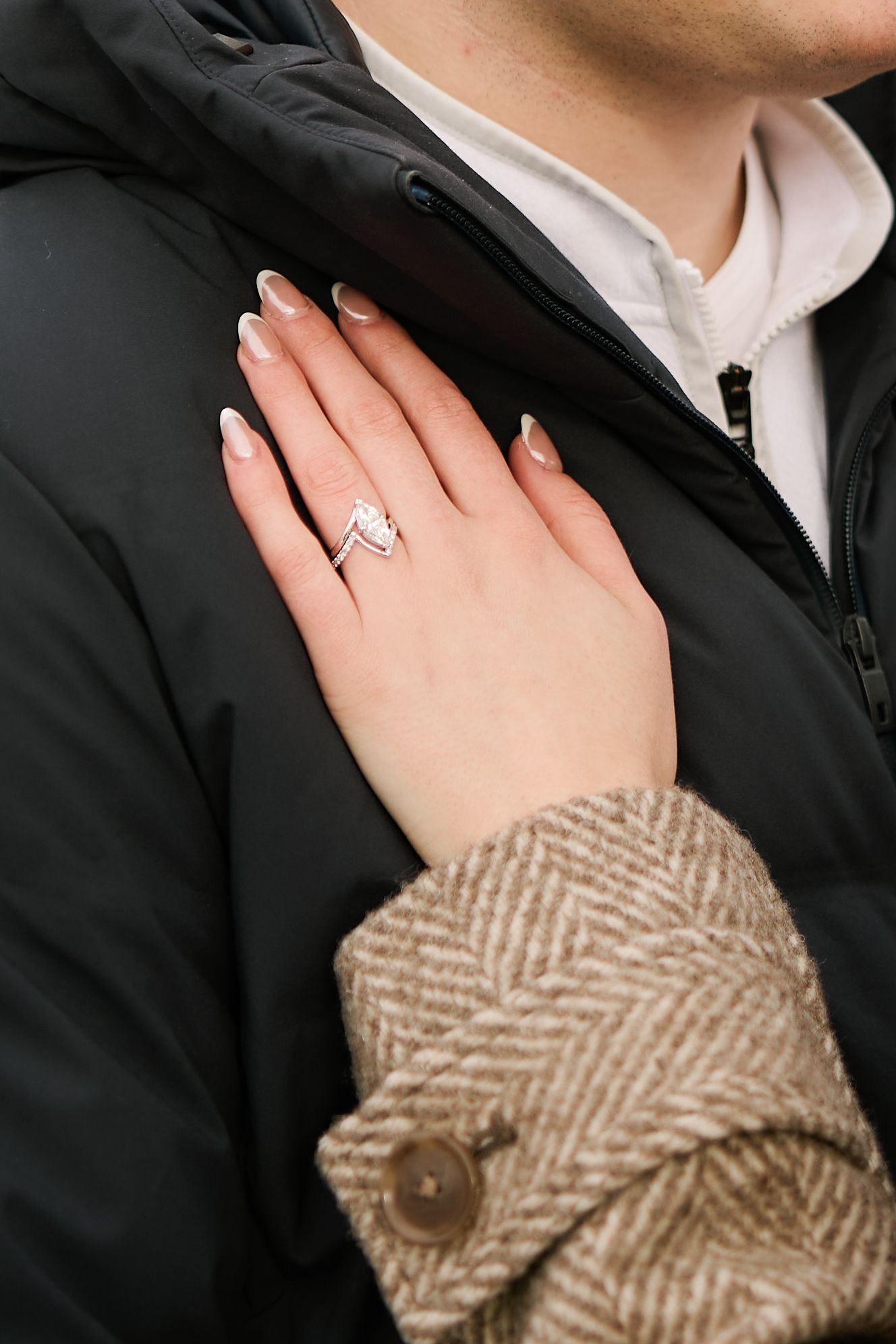 Close-up of a hand wearing a diamond engagement ring against a black winter coat and brown tweed sleeve.