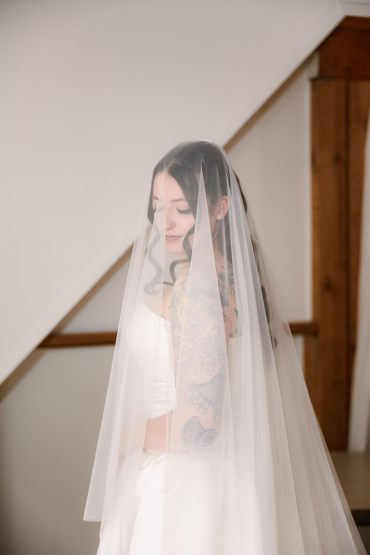 Bride in white wedding dress and flowing veil standing near staircase, with visible tattoos on her arm.