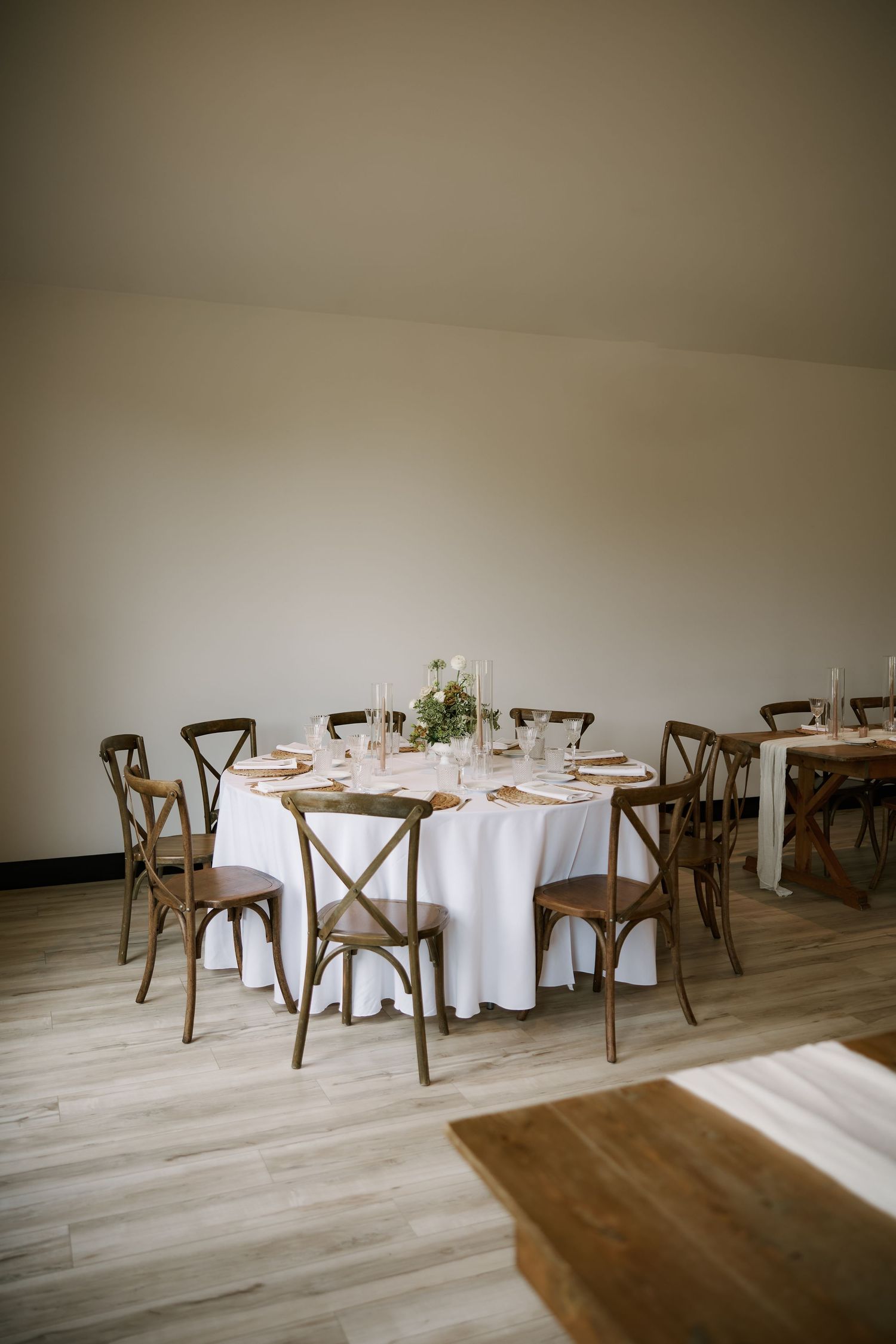 Elegant round wedding reception table with white linen, gold chargers, floral centerpiece, and rustic cross-back wooden chairs.