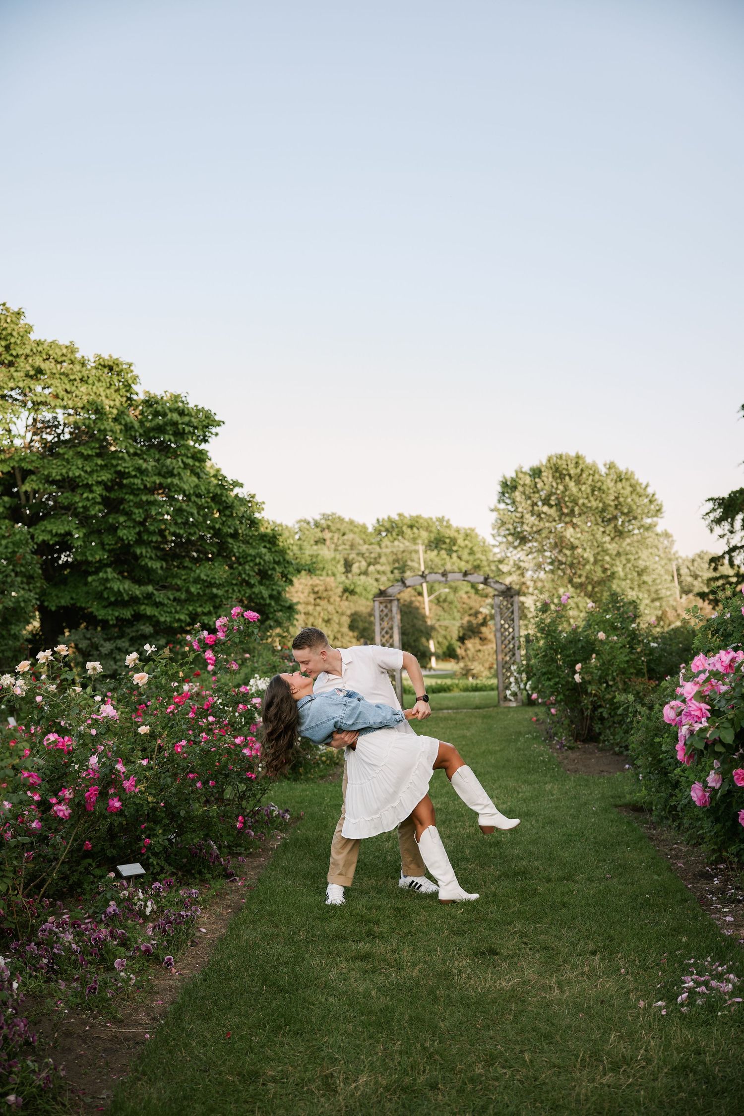 Couple sharing a romantic dip kiss in a lush garden path lined with blooming pink roses and green trees.