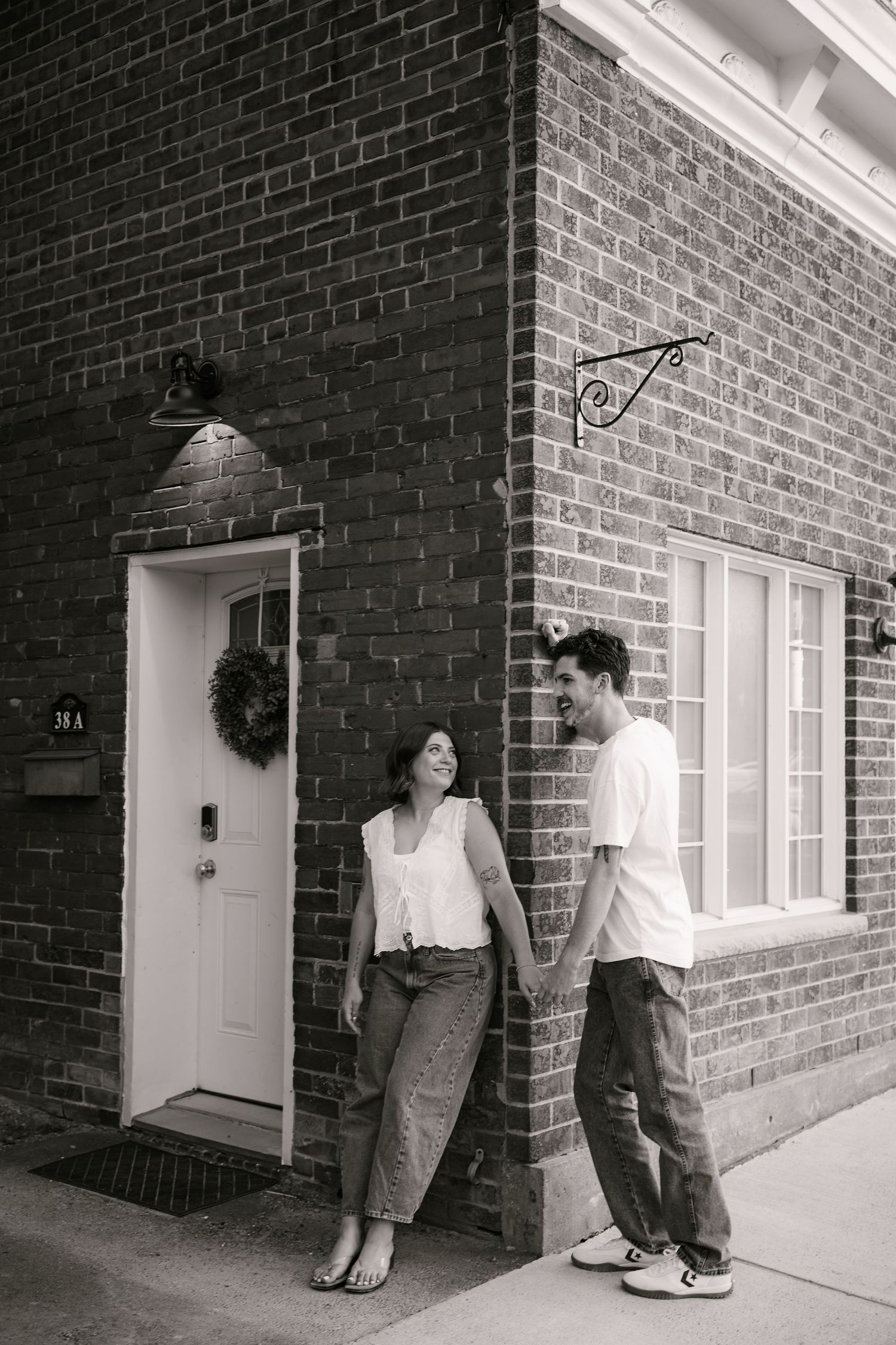 Couple smiling at each other leaning against brick building in B&W photo.