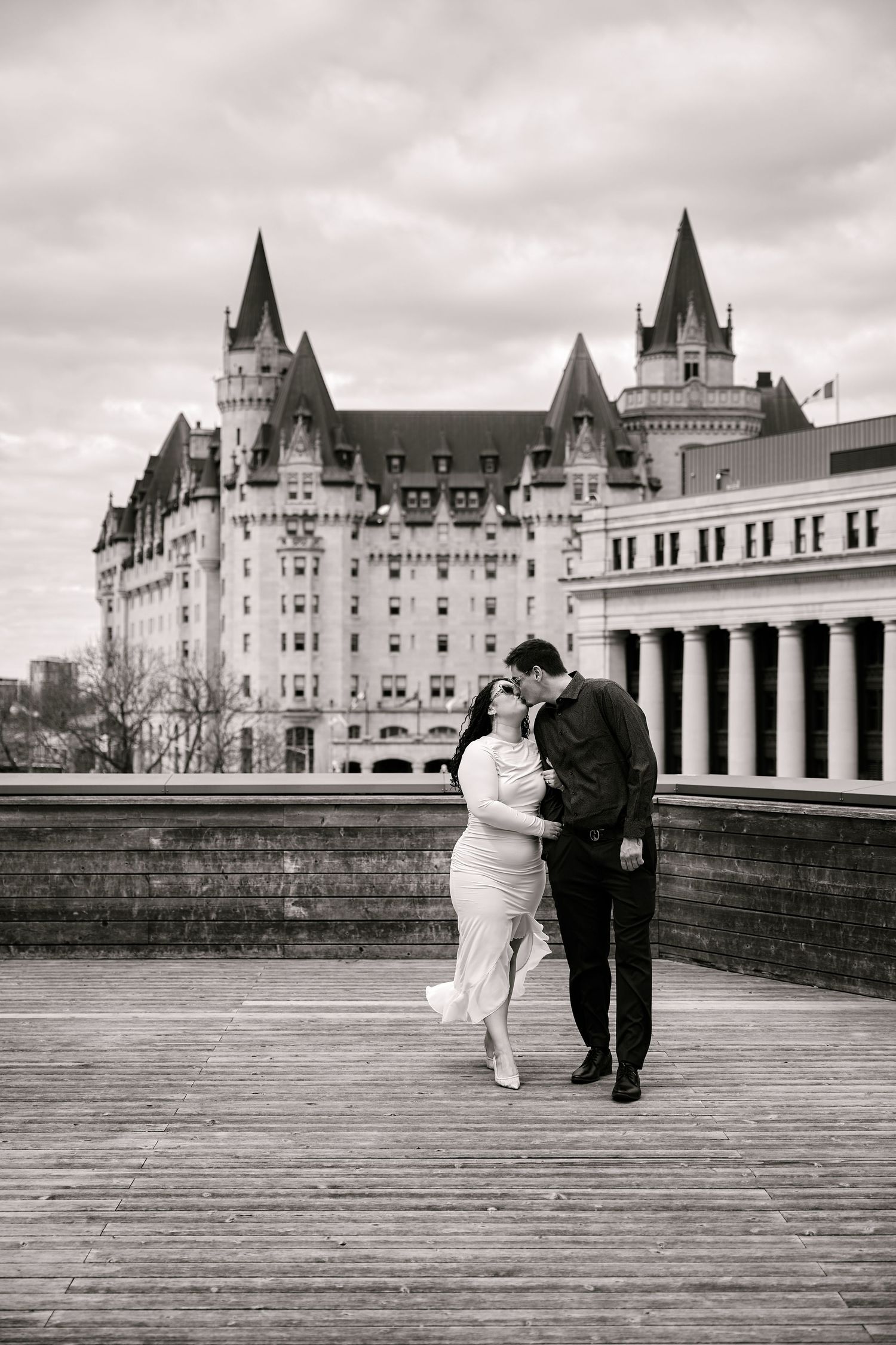 Black and white photo of a couple embracing on a wooden deck with Chateau Laurier in Ottawa behind them.