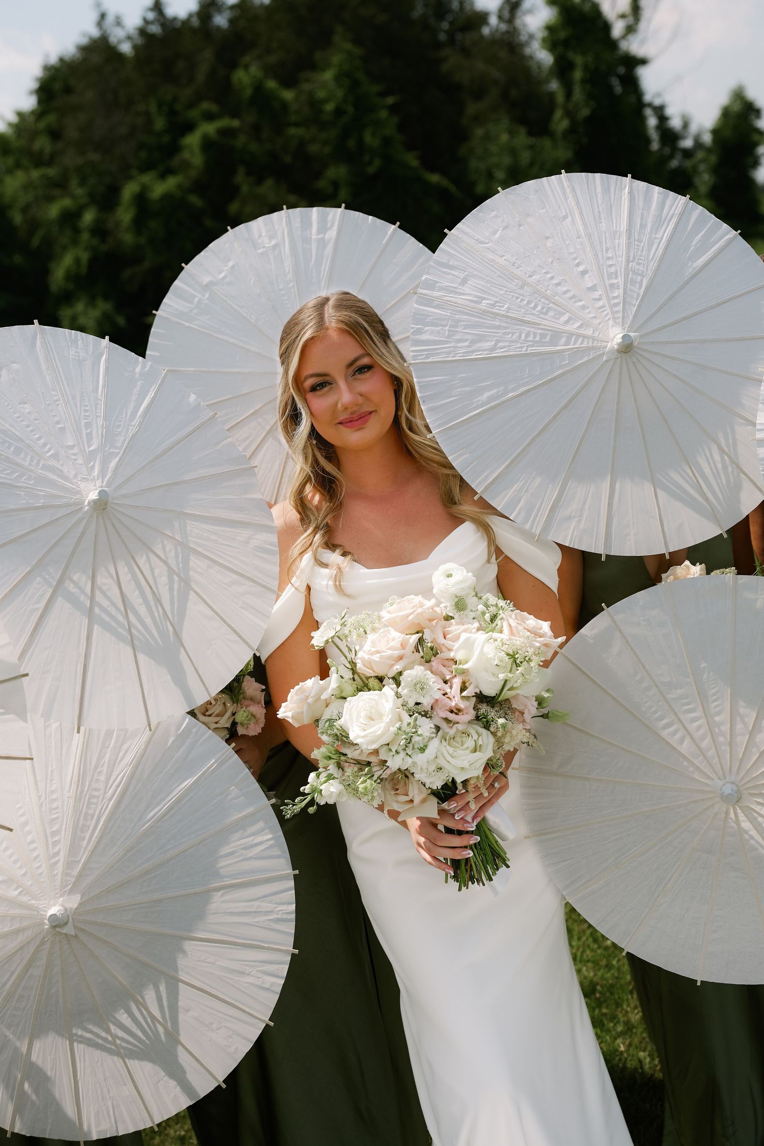 Bride in white gown holding floral bouquet surrounded by bridesmaids with white parasol umbrellas outdoors.