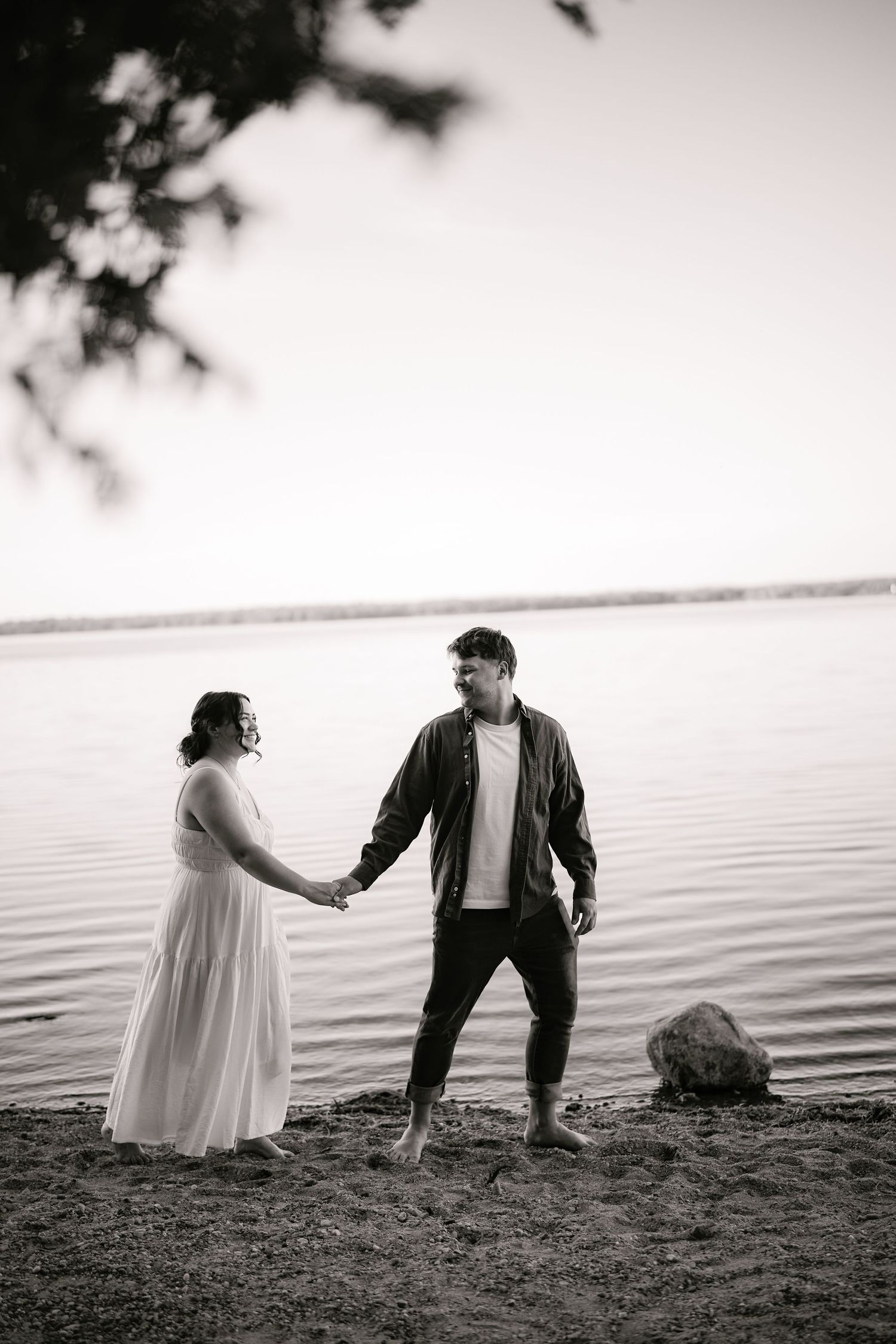 A couple holds hands while standing on the shoreline of a lake in black and white.