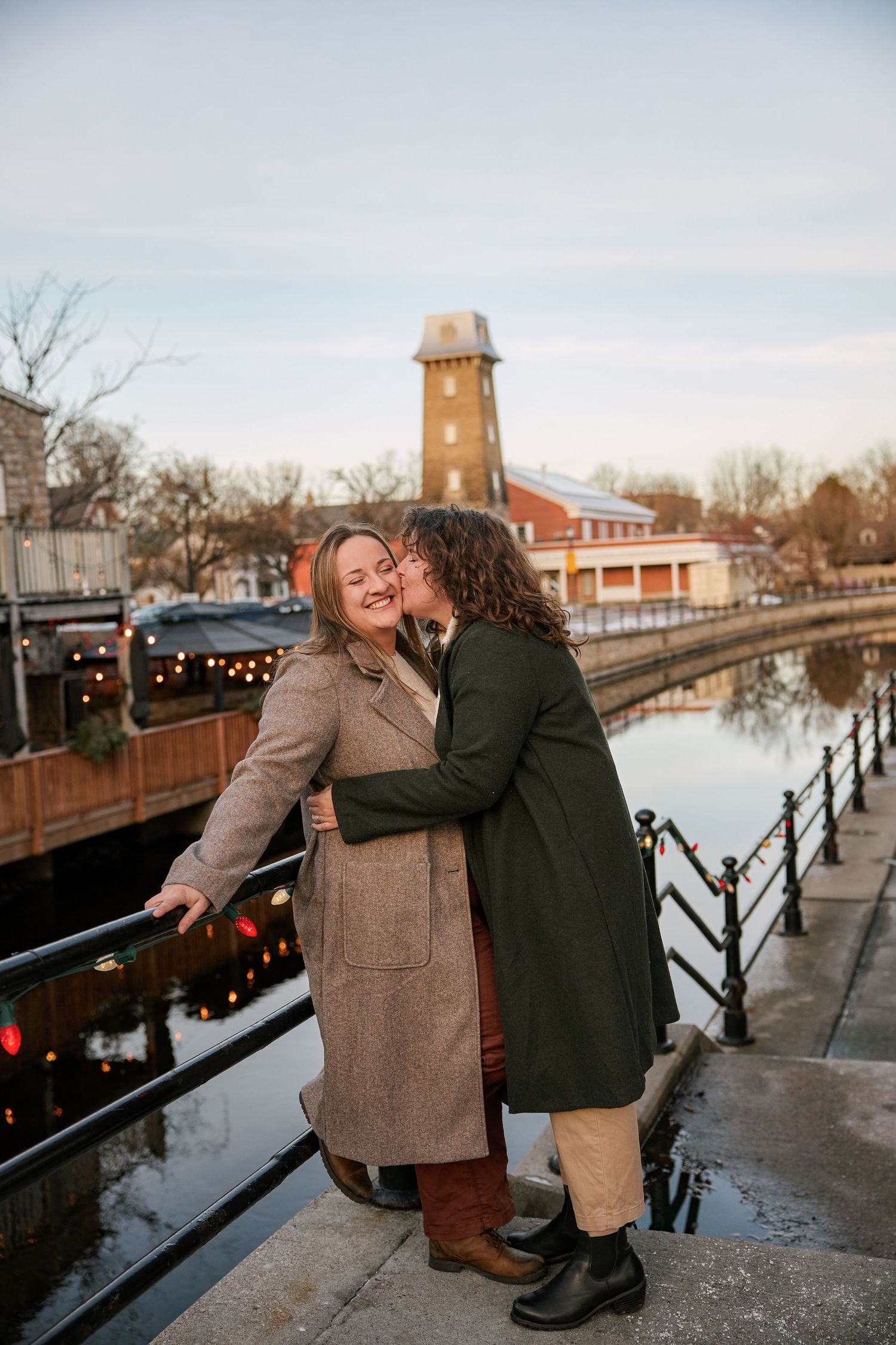 Two women sharing a romantic kiss on a canal bridge in winter coats, with a historic watermill and string lights in background.