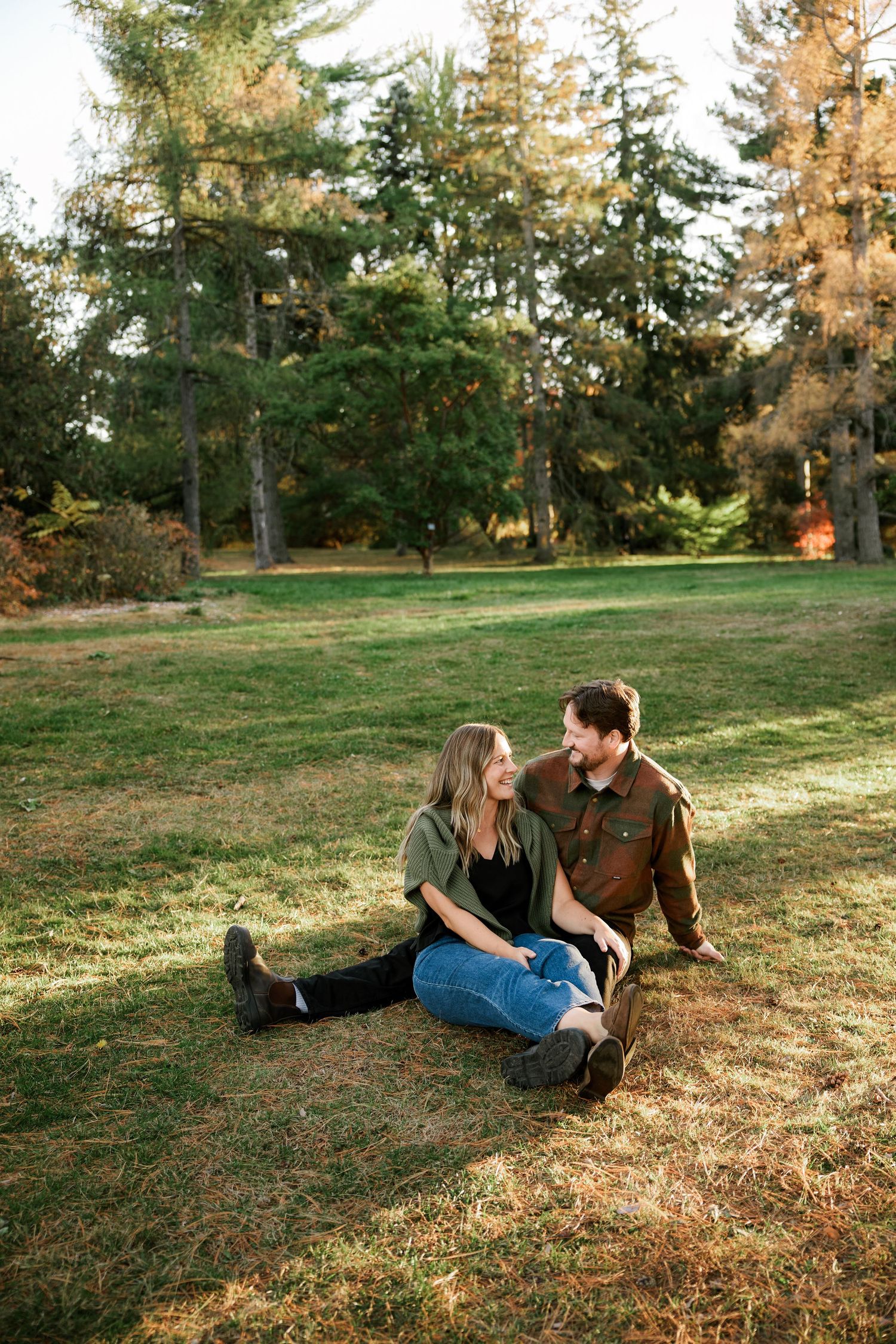 Couple sitting together on grass in a park surrounded by tall pine trees during golden hour.