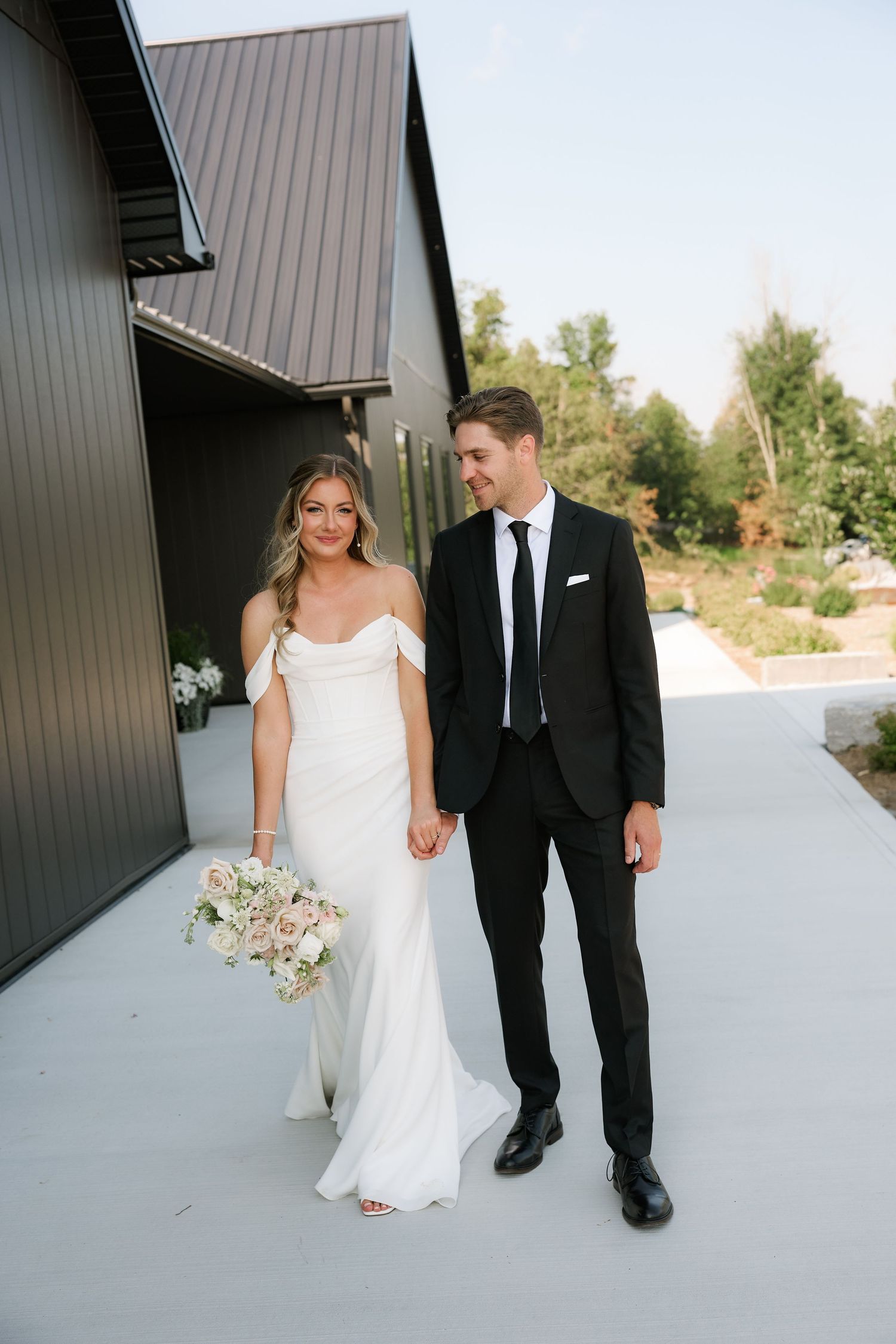 A bride in a white off-shoulder gown holds a floral bouquet beside a groom in a black tuxedo outside a modern venue.