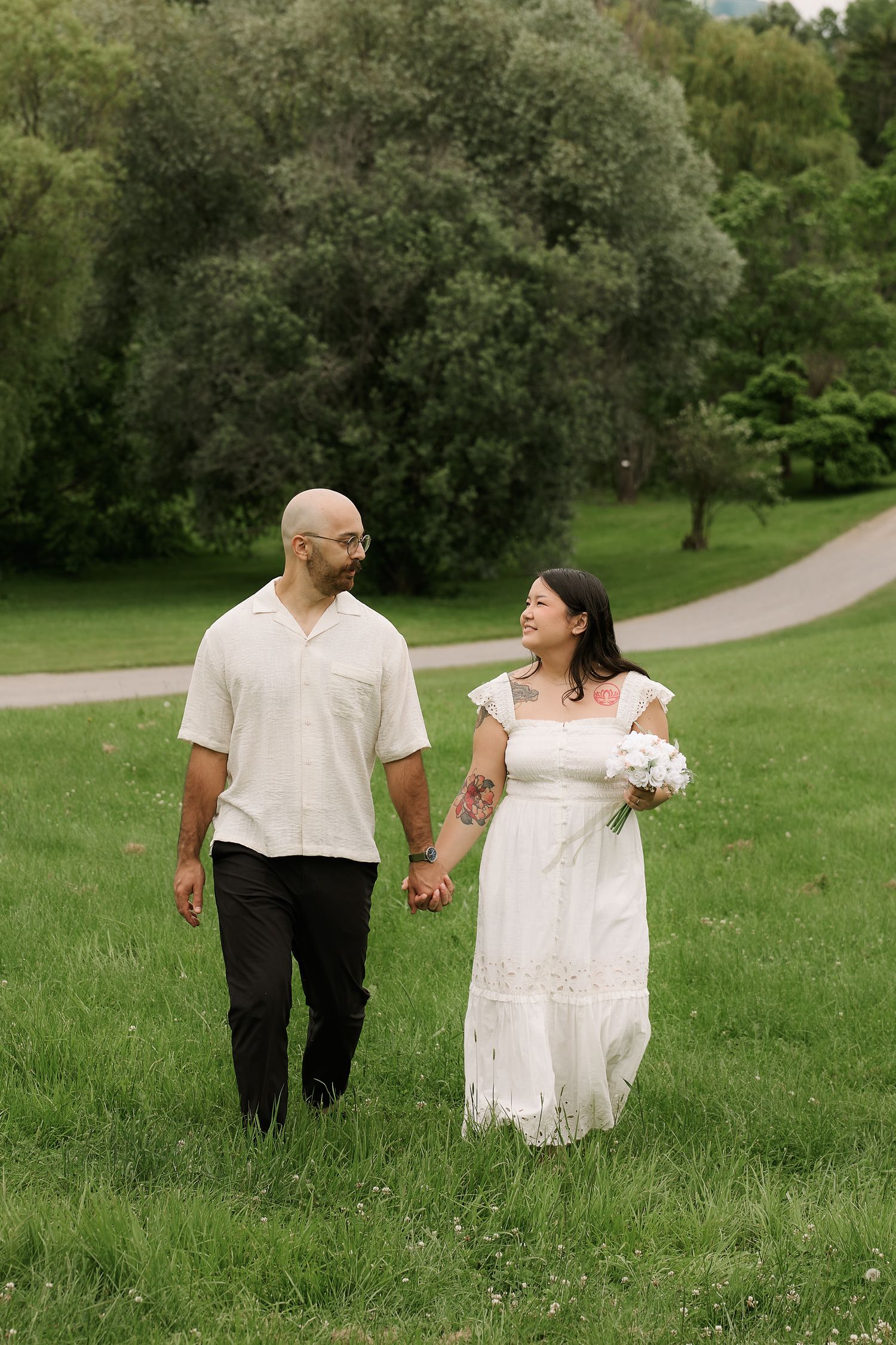 A couple in wedding attire walks hand-in-hand through a lush green park, smiling at each other.