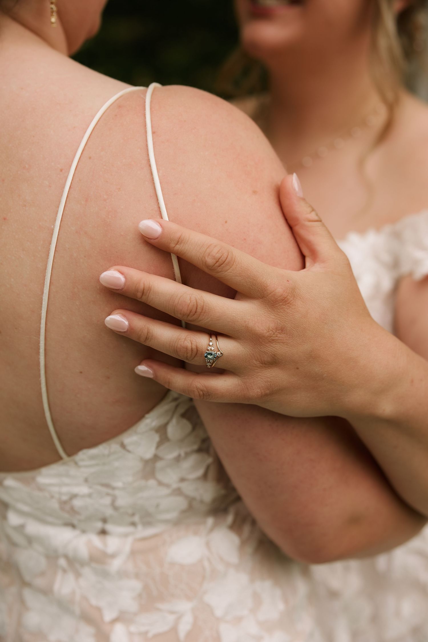 Close-up of two brides embracing, showcasing a delicate diamond engagement ring and soft pink manicure against a white lace wedding dress.