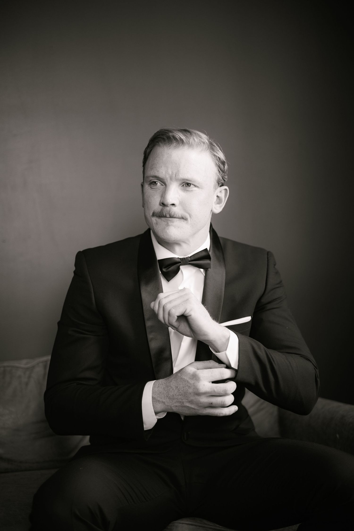 Black and white portrait of a mustachioed man in a tuxedo holding a cigarette and glass, seated against a dark background.