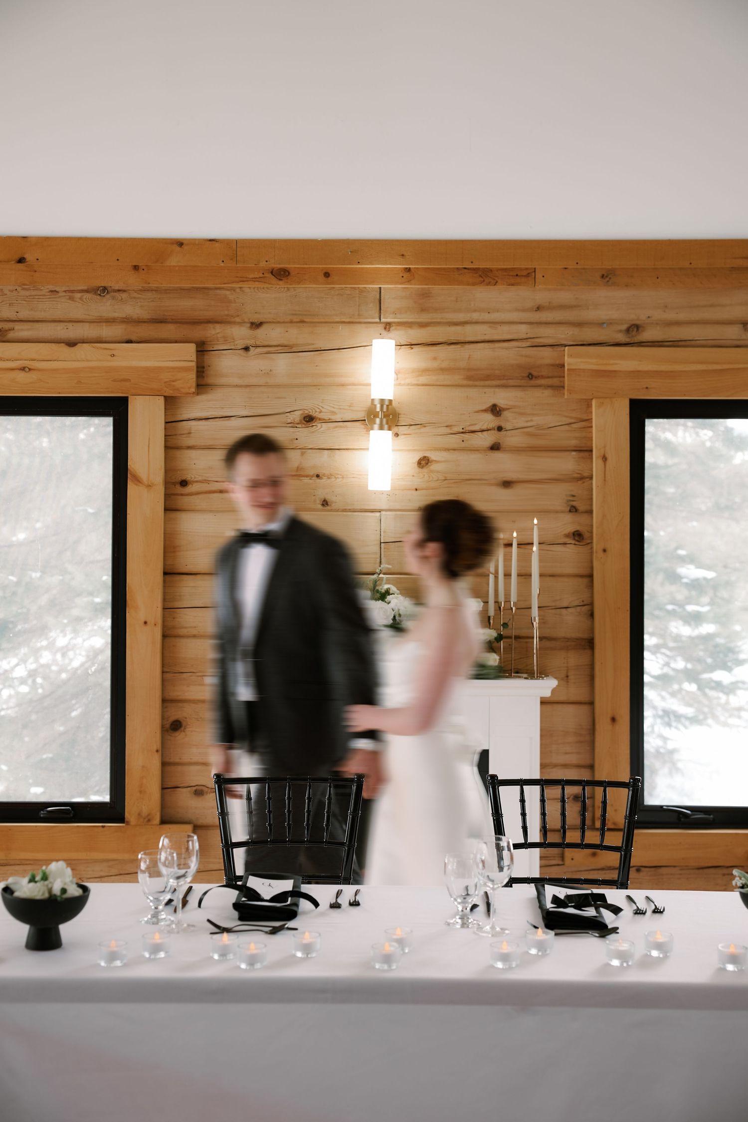 Motion-blurred shot of wedding couple at reception table with rustic wood walls and windows.
