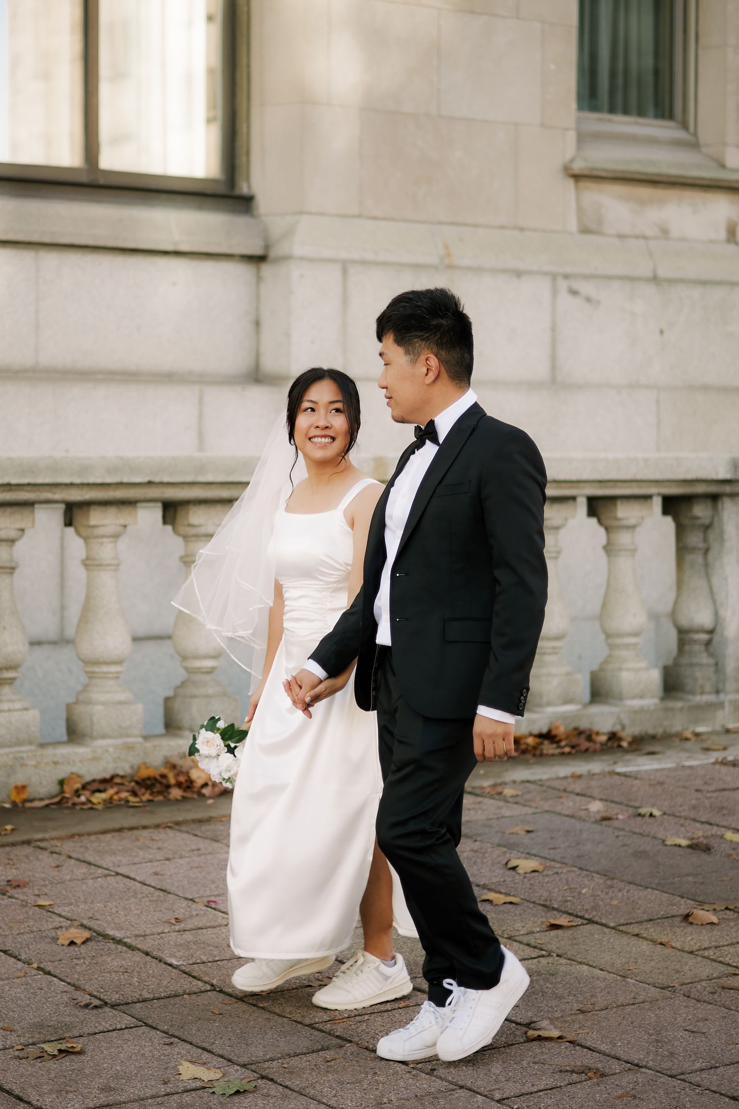 Asian bride in white dress and veil holds bouquet while walking with groom in black tuxedo on stone pathway.