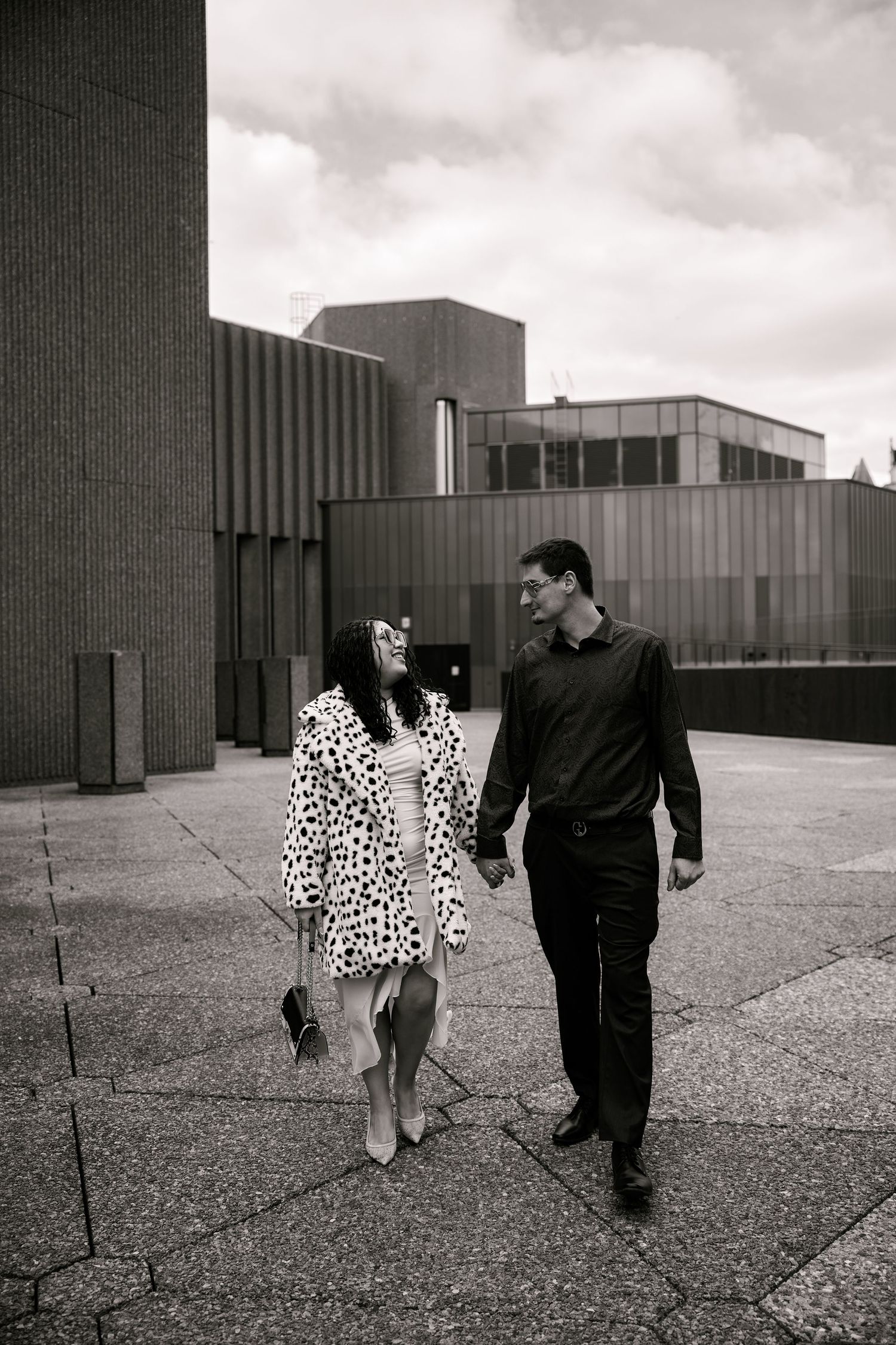 Black and white photo of a couple holding hands and walking together outdoors near modern buildings.
