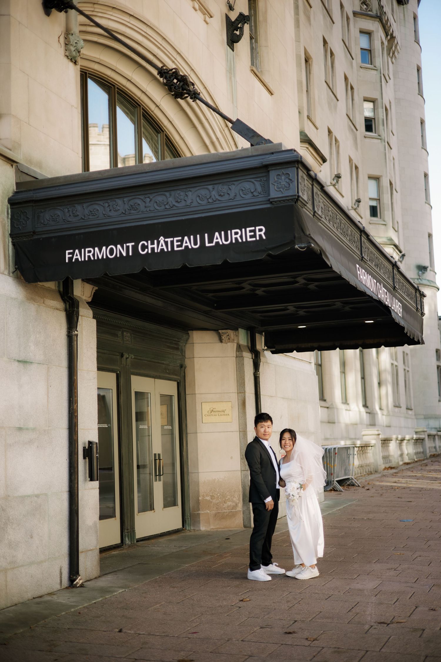 Couple in wedding attire posing outside Fairmont Château Laurier hotel entrance on a cobblestone sidewalk.