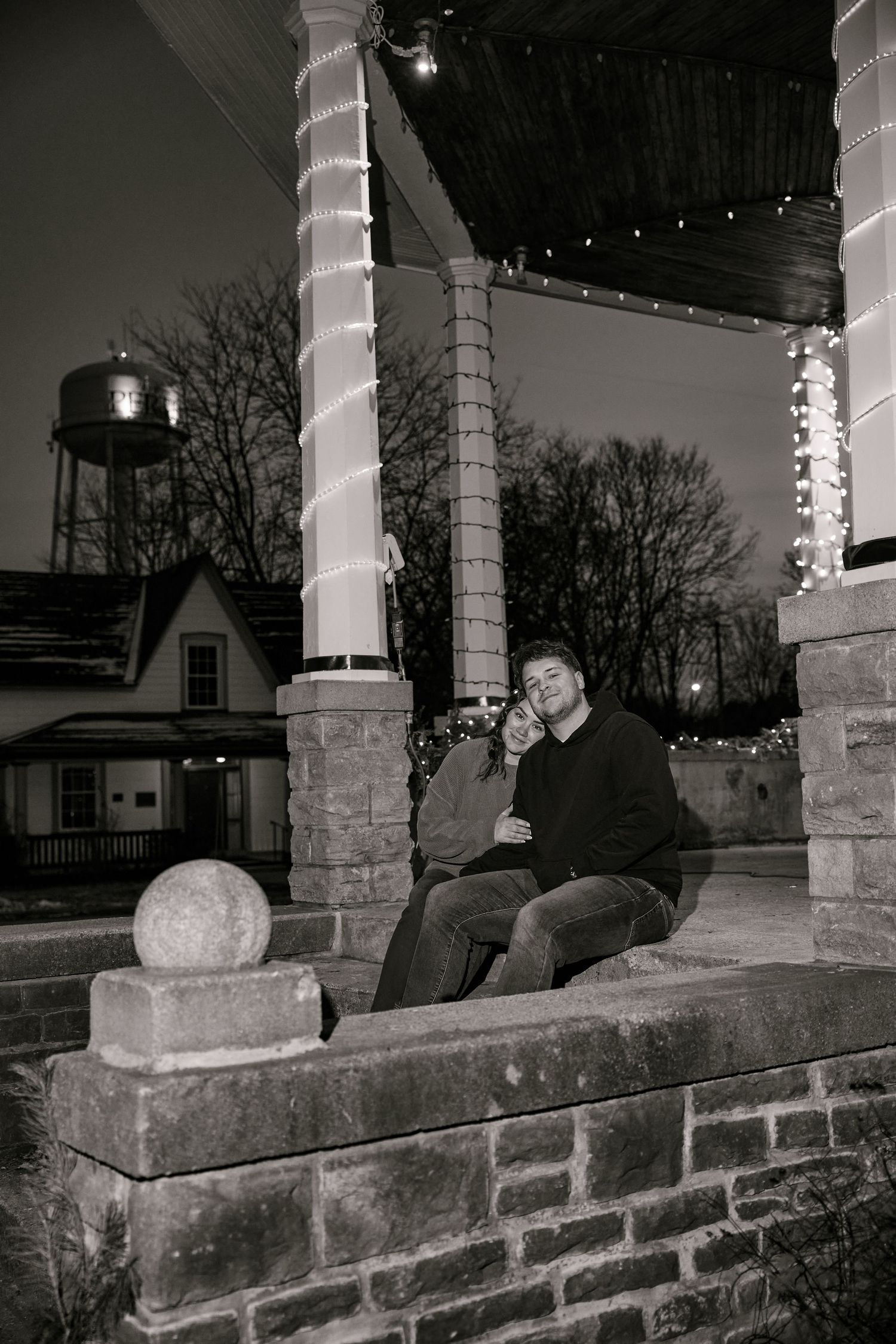 Man sitting on stone steps of a gazebo decorated with Christmas lights at night, in black and white photo.