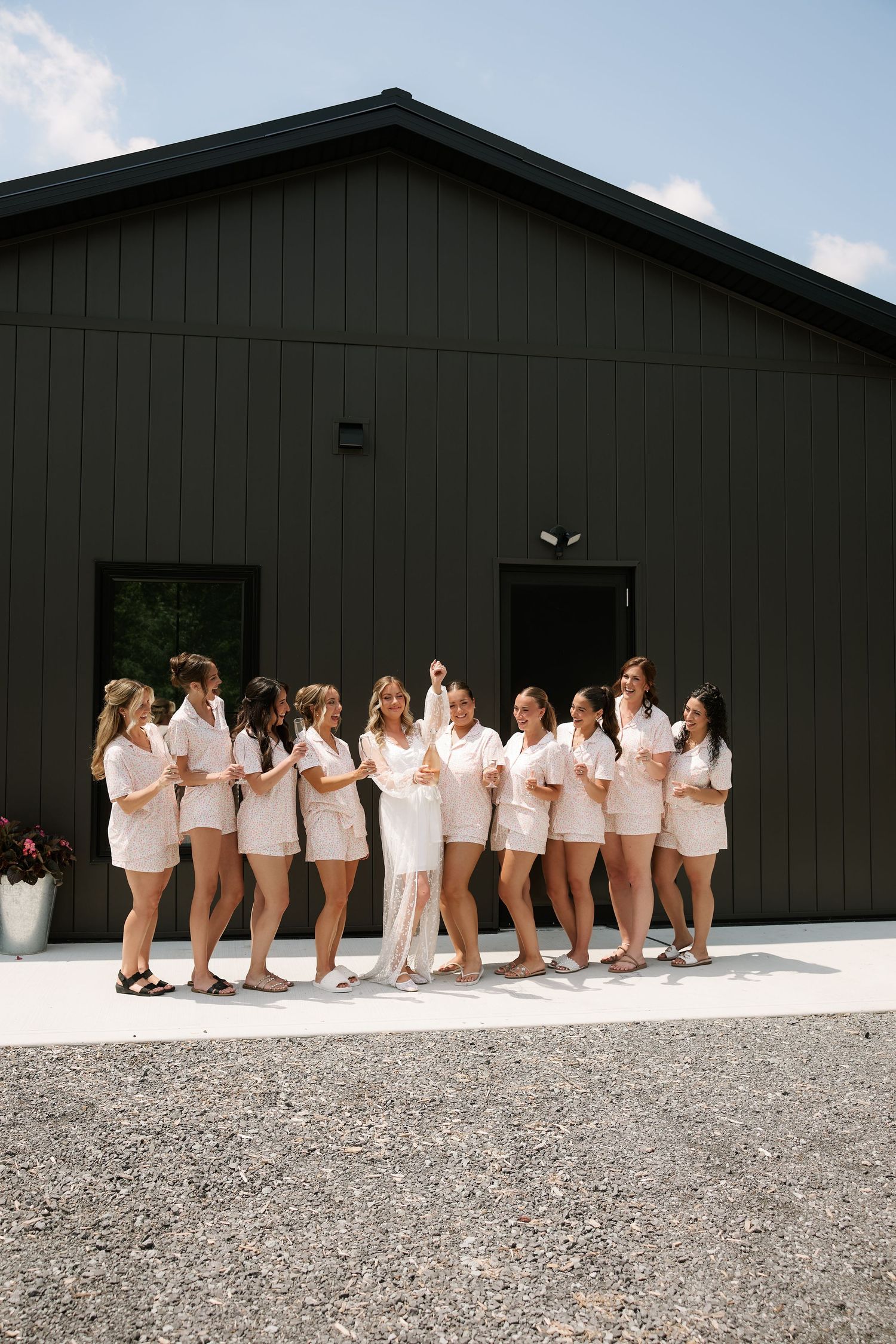 Bride in white pantsuit poses with bridesmaids in matching pink sets outside a dark barn venue.