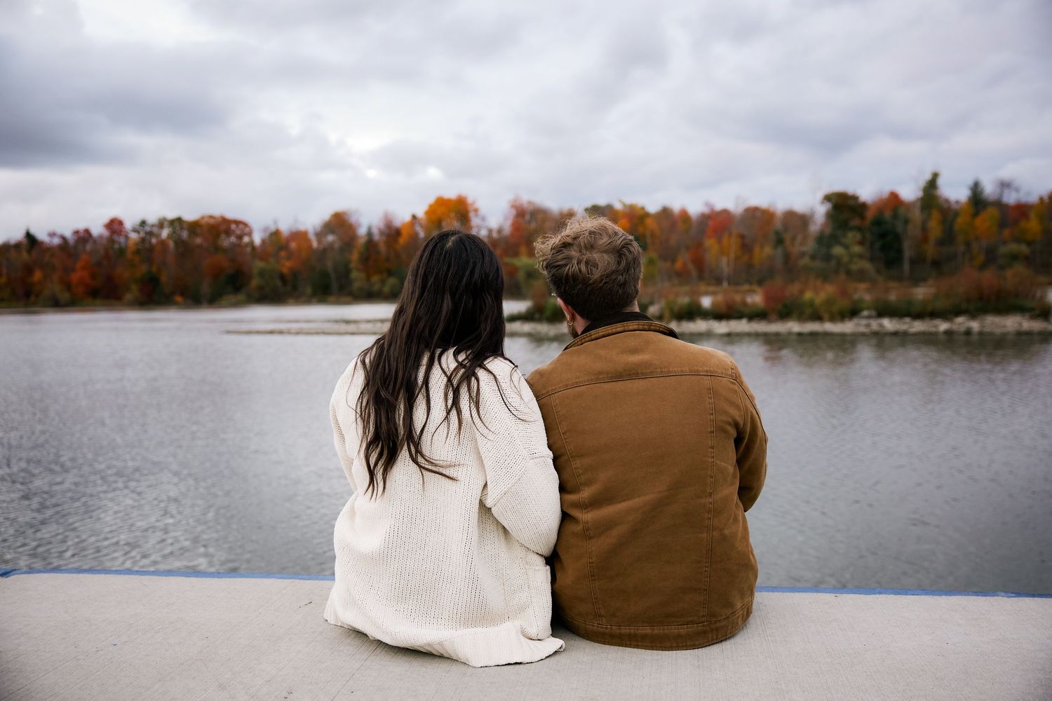 Couple sitting together on a ledge overlooking a calm lake surrounded by colorful autumn foliage.