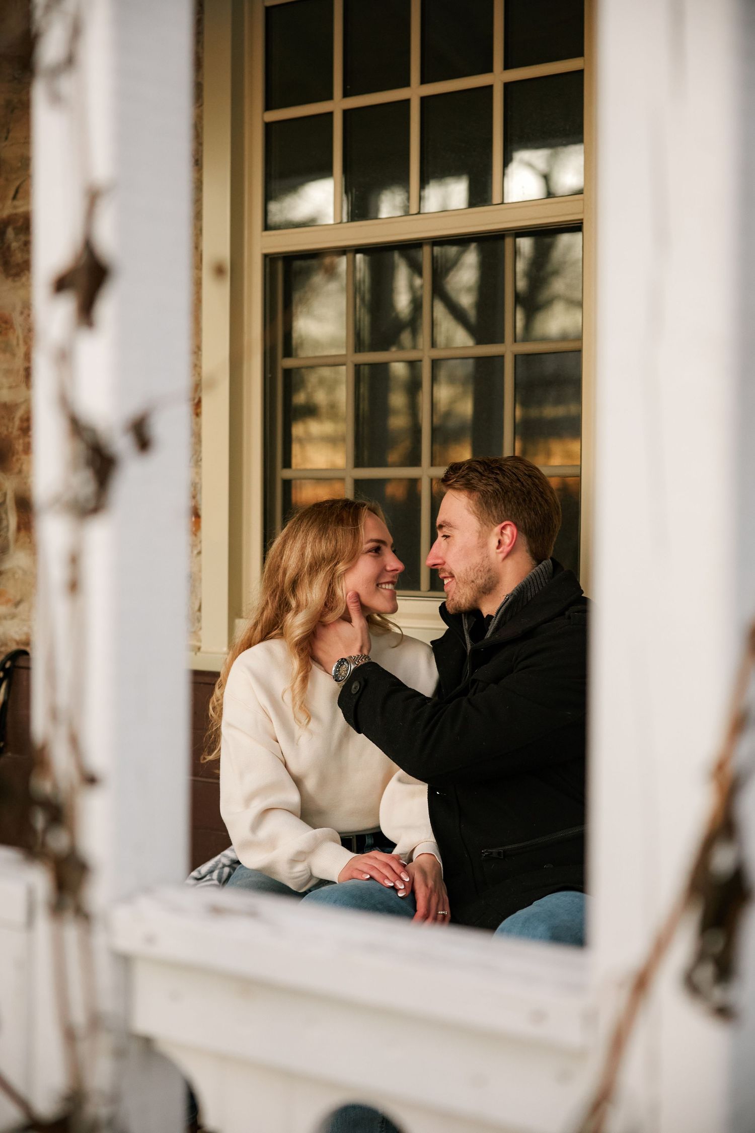 A smiling couple sits together on a porch windowsill, gazing at each other affectionately in casual winter attire.
