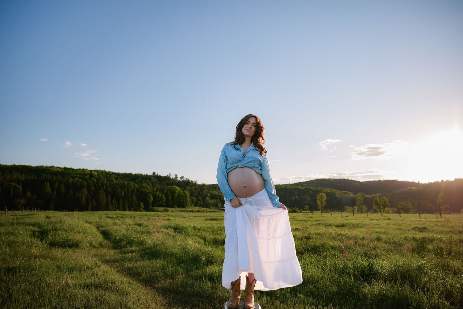 A figure in a white skirt and blue top stands in a sunlit meadow at golden hour.