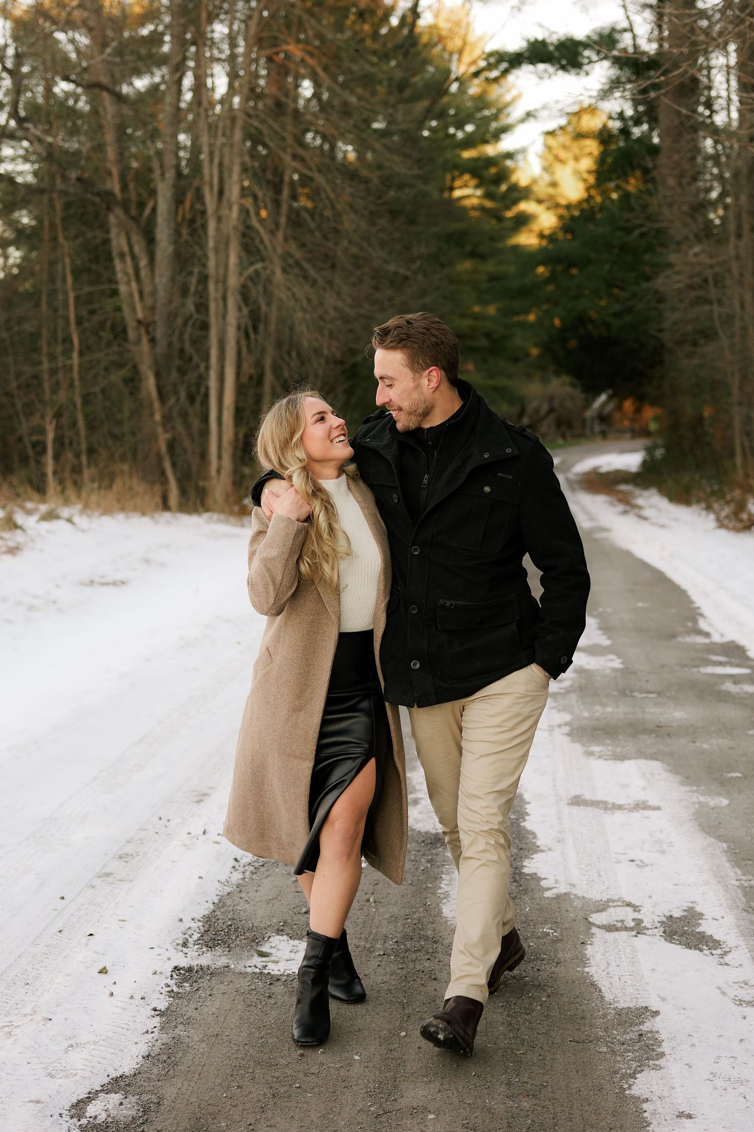 A couple walking together on a snowy path through a winter forest at sunset.