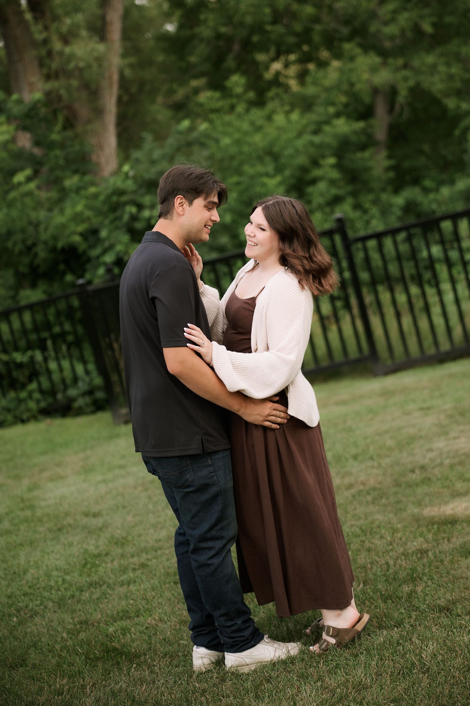 A smiling couple embraces on a green lawn, the woman in a brown dress and white cardigan, the man in a black shirt and jeans.