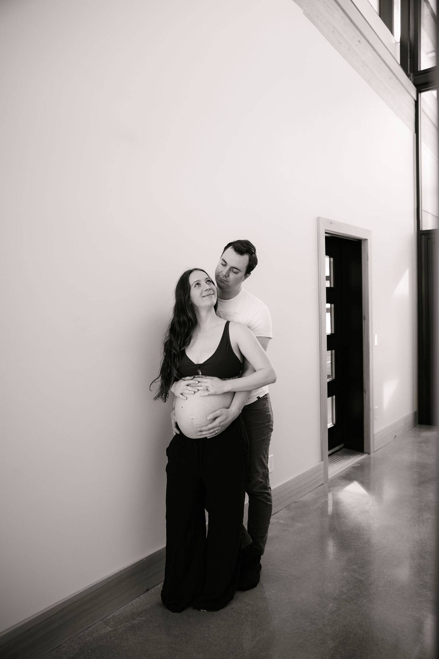 A black and white maternity photo in a minimalist hallway setting.